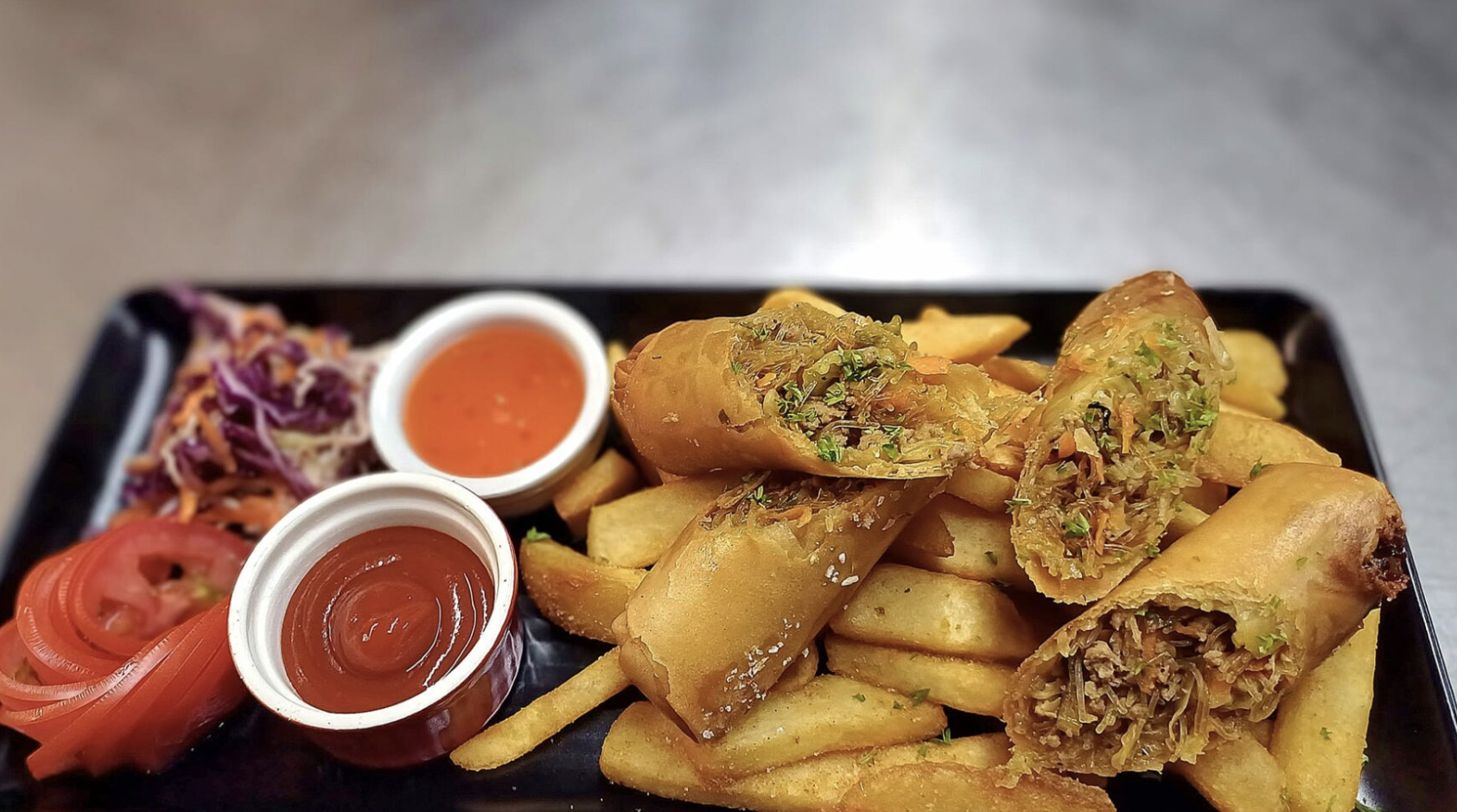 Loaded spring rolls with dipping sauces, fresh sliced tomatoes, coleslaw, and crispy fries on a black rectangular plate.
