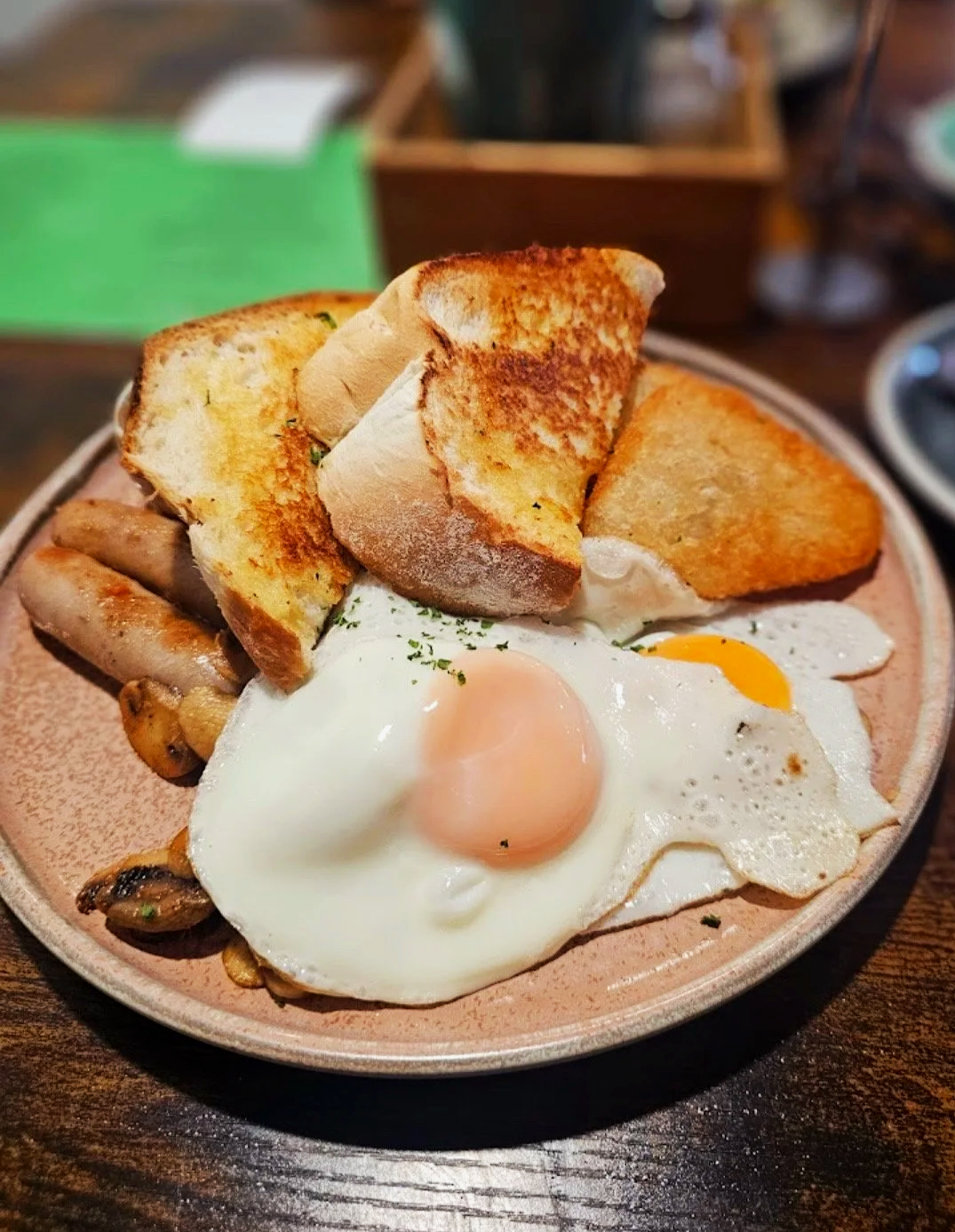 A plate of breakfast food with fried eggs, toasted bread, sausages, and roasted potatoes on a ceramic plate from stone oven bakery and cafe in Hokitika 