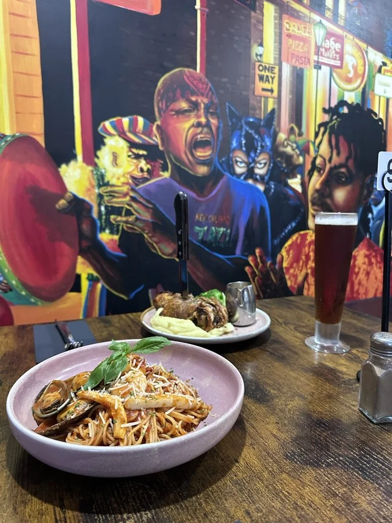 A plate of seafood pasta garnished with basil, a plate with ribs, mashed potatoes, and a drink on a wooden table in a restaurant with a colorful mural wall.