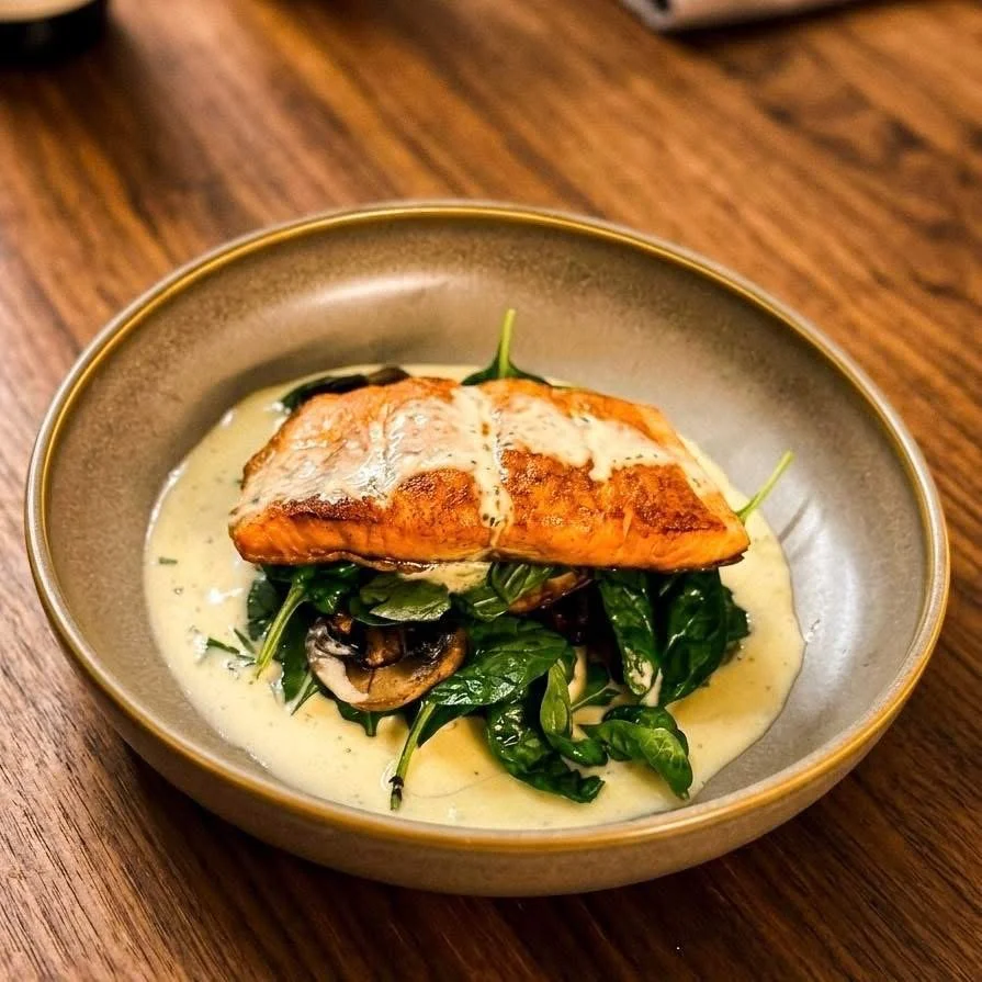 A plated meal featuring a piece of cooked fish, possibly salmon, on a bed of cooked leafy greens and mushrooms, with creamy sauce drizzled over the fish, served in a shallow bowl on a wooden table.