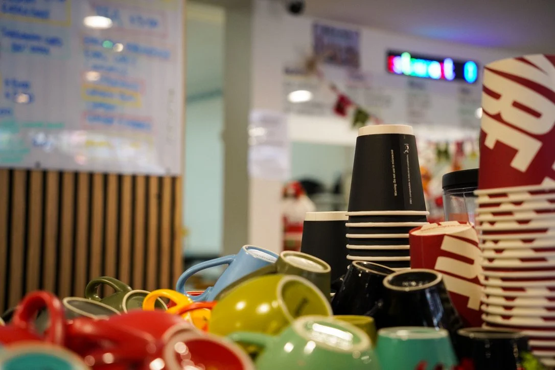 Stacked disposable coffee cups, colorful ceramic mugs, and a red and white striped paper bag on a counter at a coffee shop.