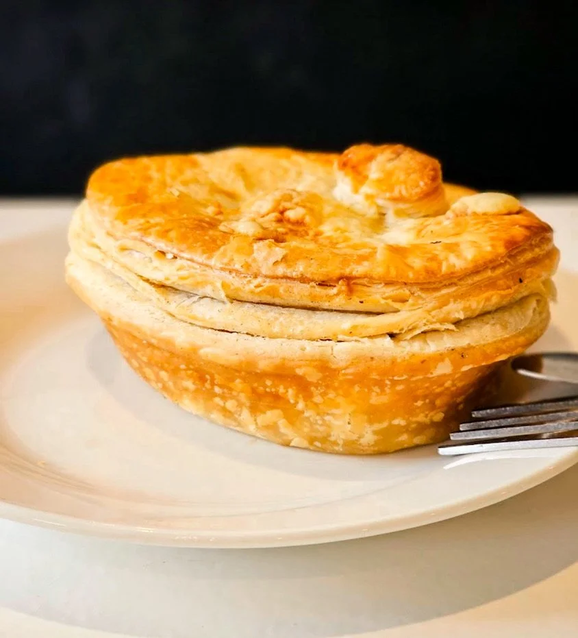 A plate with a folded, golden-brown, flaky pastry or bread topped with a few small pieces of dough or crumbs. A fork is placed beside the plate.