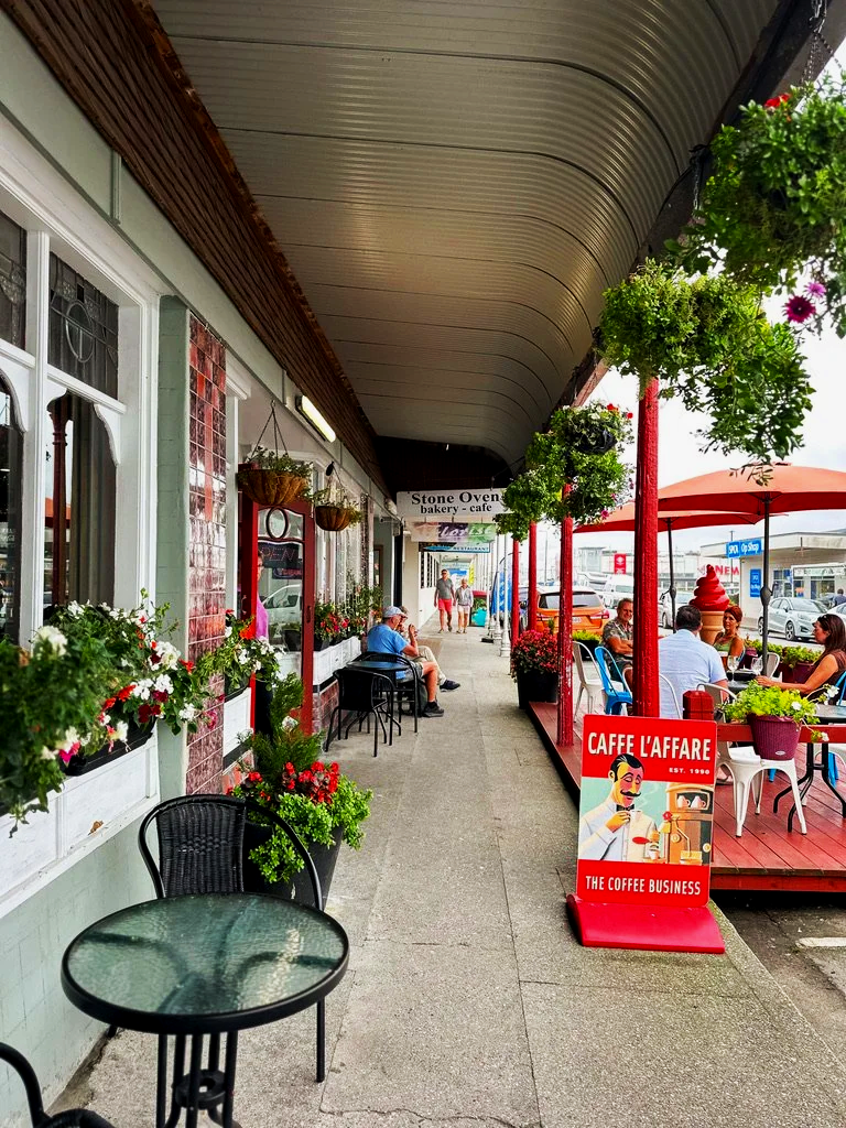 Sidewalk cafe with outdoor seating under a covered awning, decorated with hanging plants and flowers, with a restaurant sign that reads "Stone Oven bakery - cafe", and a red sign advertising "Caffe L'Affare the coffee business" near a group of people dining outside.