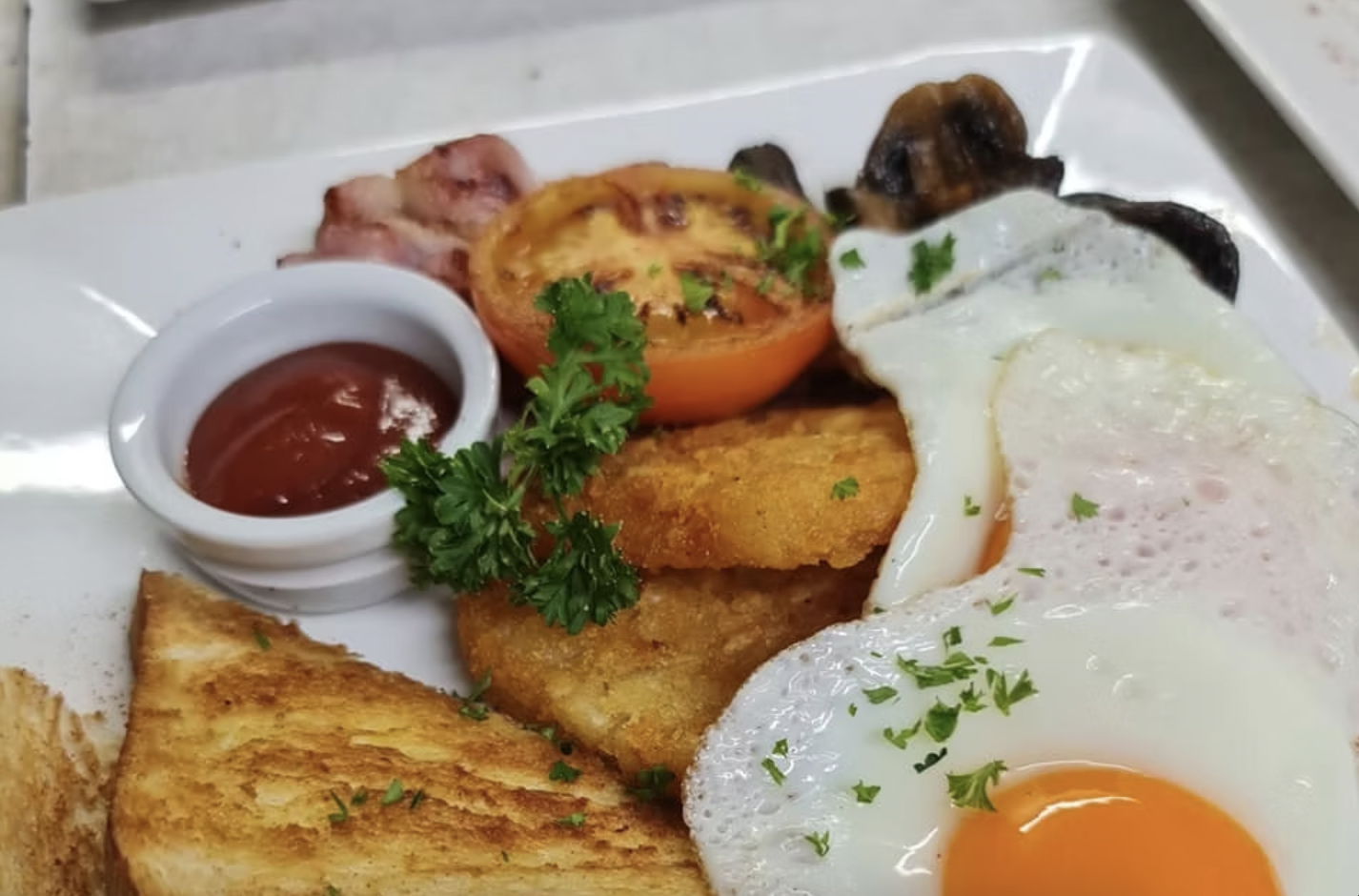 Plate of breakfast food including sunny side up eggs, toast, tomatoes, hash browns, bacon, sausage, mushrooms, and ketchup garnished with parsley.