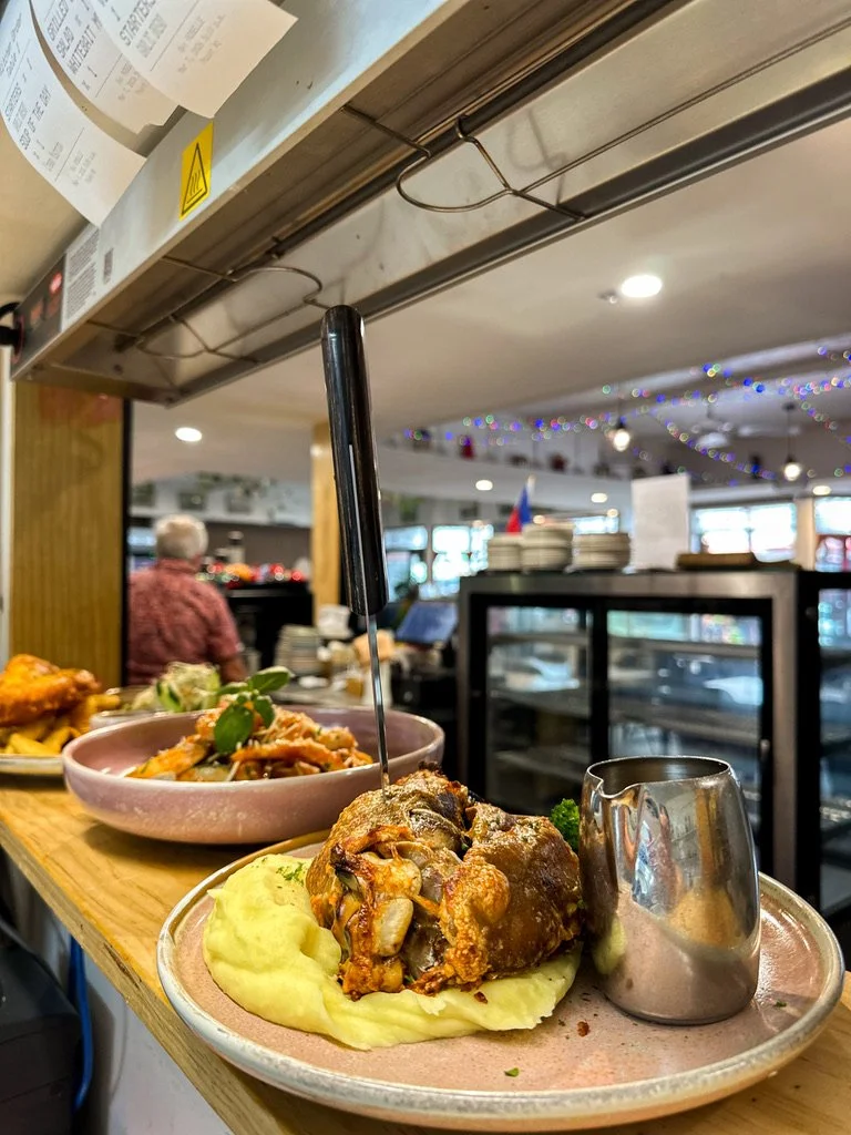 A plate of fried meat with mashed potatoes and gravy served at a restaurant. In the background, there are other dishes, a person wearing a red patterned shirt, and festive decorations.
