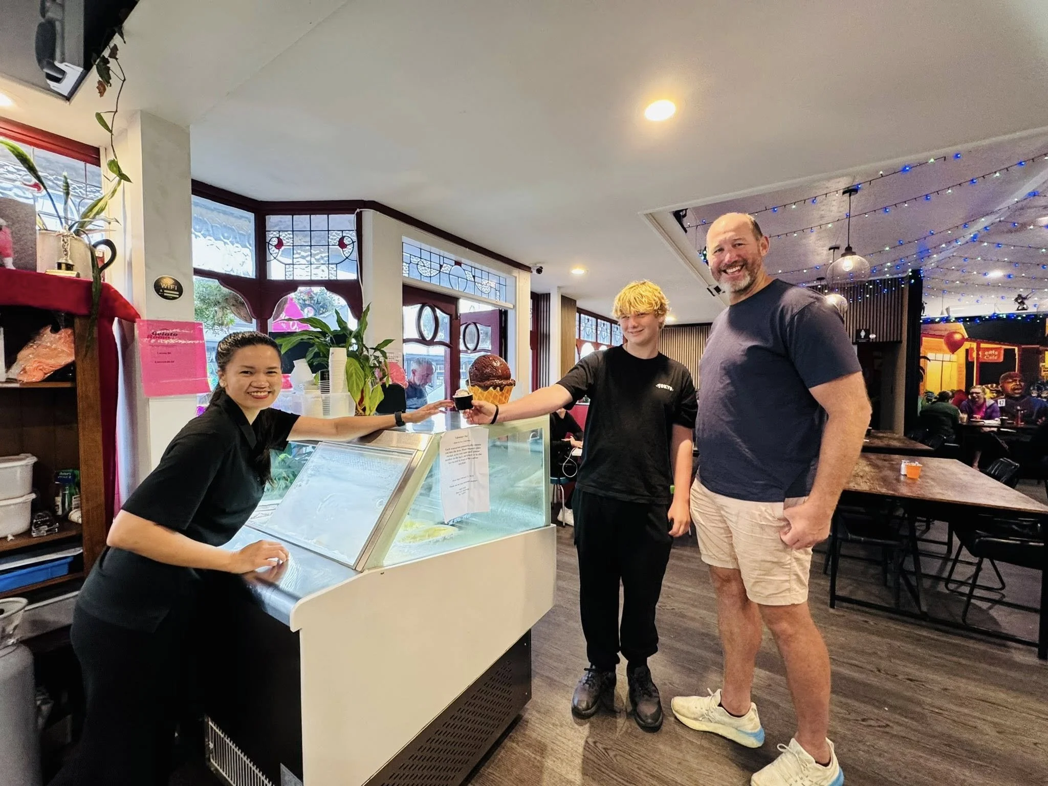 A woman serving ice cream to a customer at an ice cream shop. The customer is receiving a large ice cream cone with two scoops. There are two people, a woman and a man, smiling and standing inside a brightly lit restaurant decorated with string light