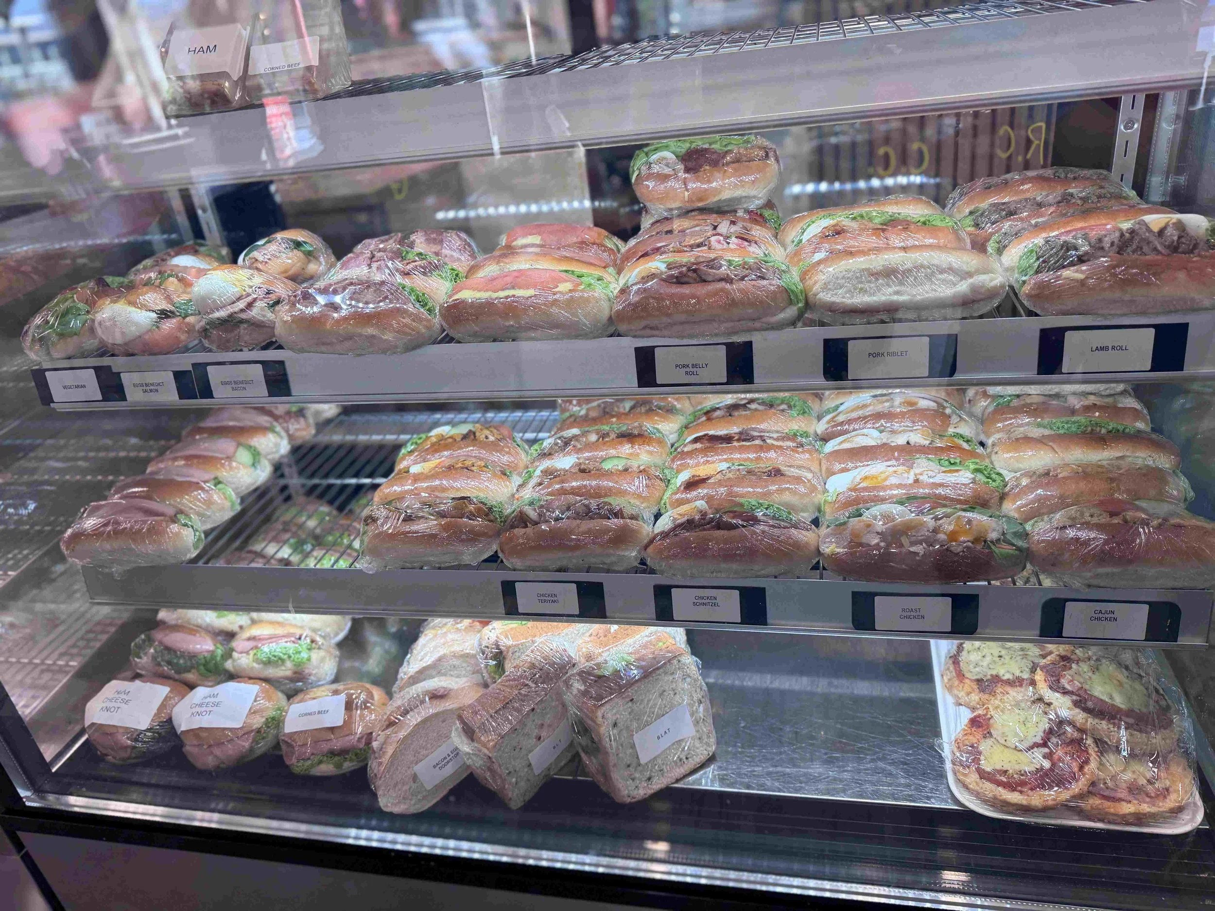 Display case with various sandwiches, including ham, pork, chicken, and cheese sandwiches, and a tray of pizza.
