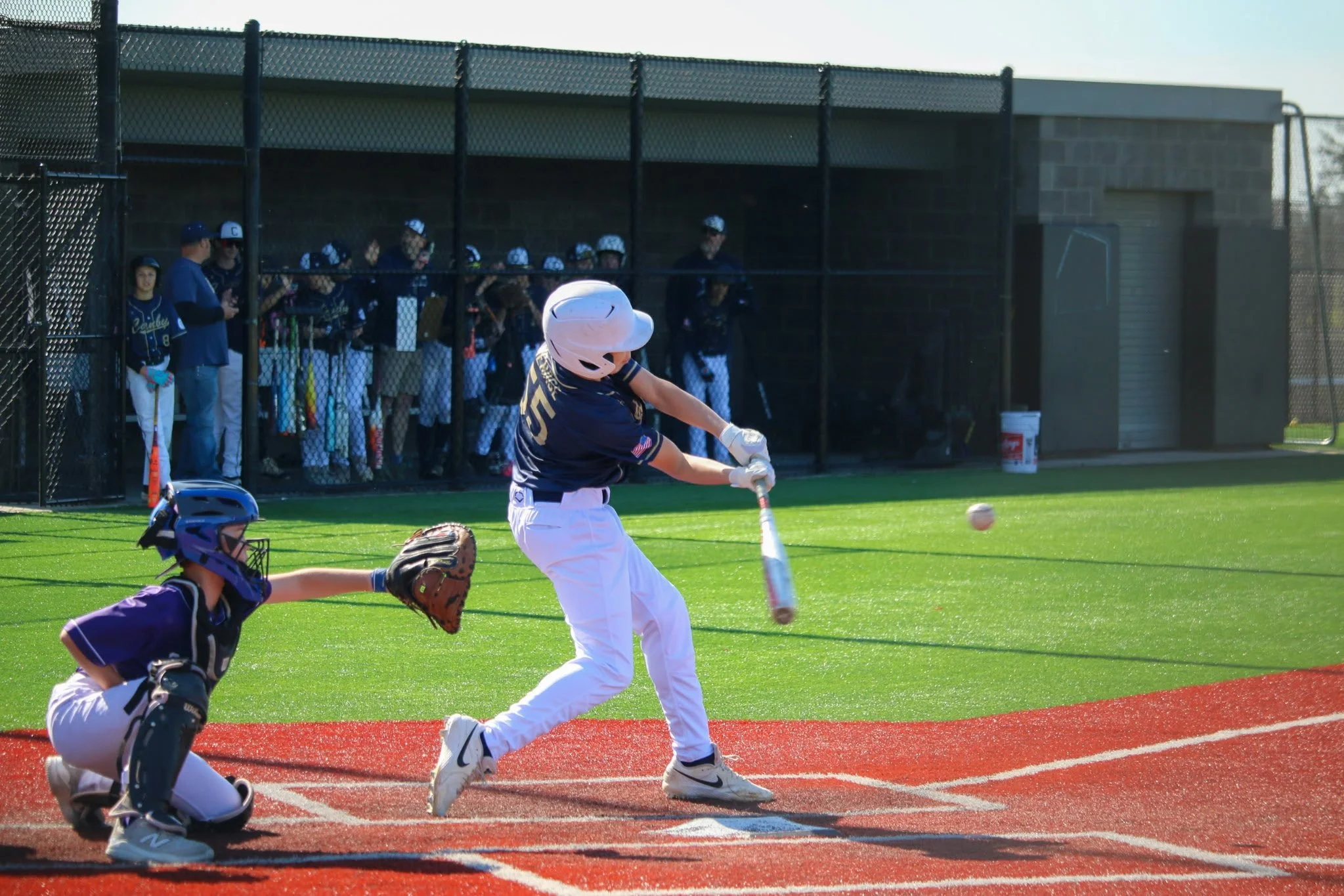 A youth baseball game on a field with a batter swinging at a pitch, a catcher on the ground, and players in the dugout watching.