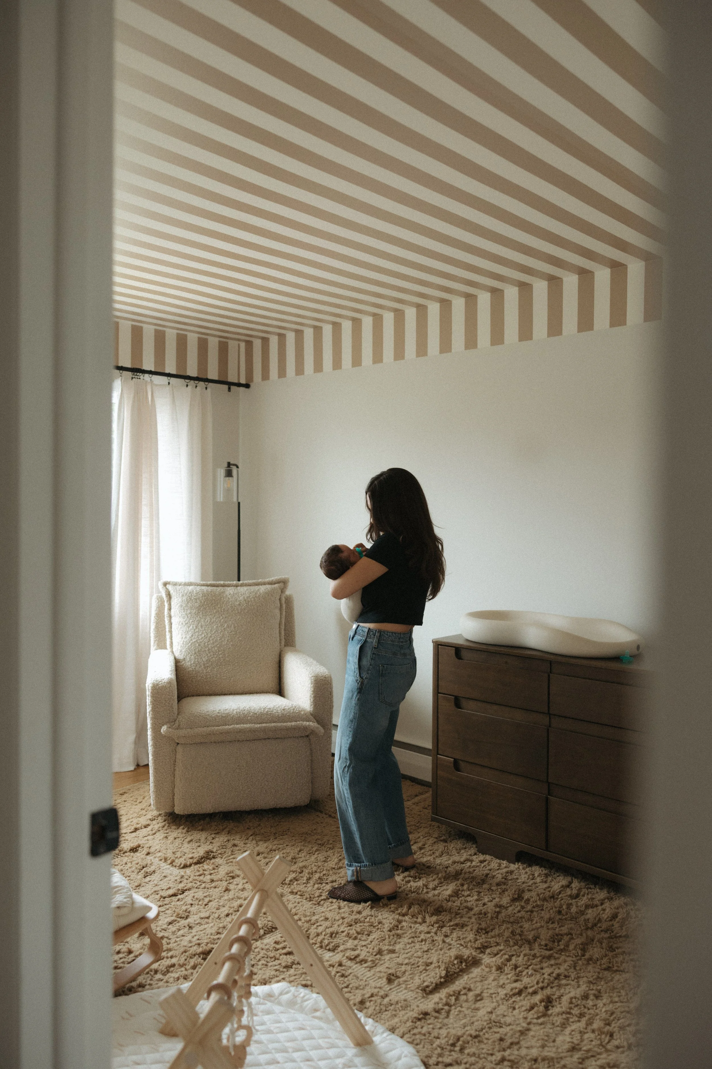 A woman holding a small baby in a nursery room with a striped ceiling, beige armchair, wooden dresser with a changing pad on top, a white curtain on a window, and a beige carpet.