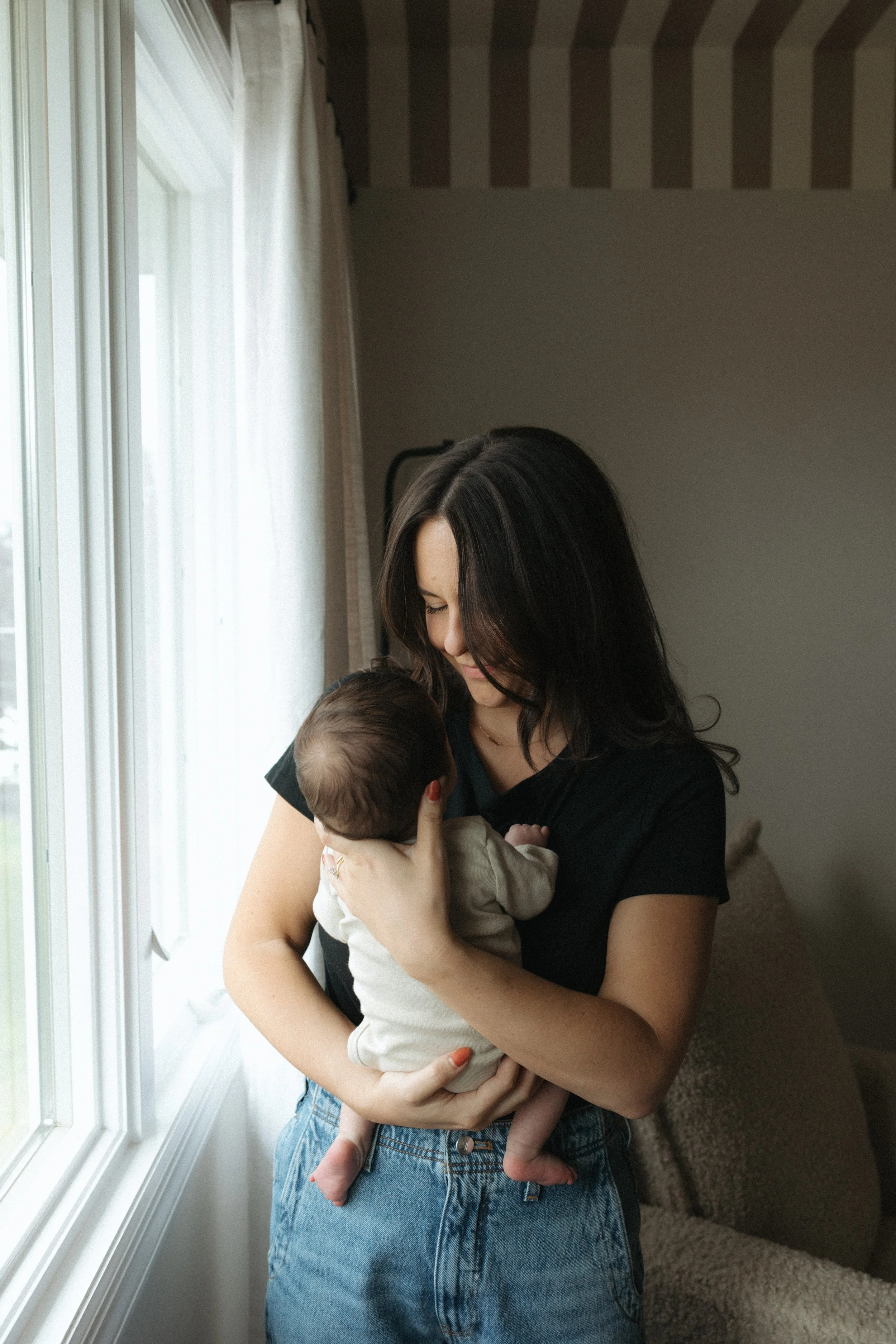 A woman with long dark hair holding a baby near a window with white curtains, interior of a home in the background.