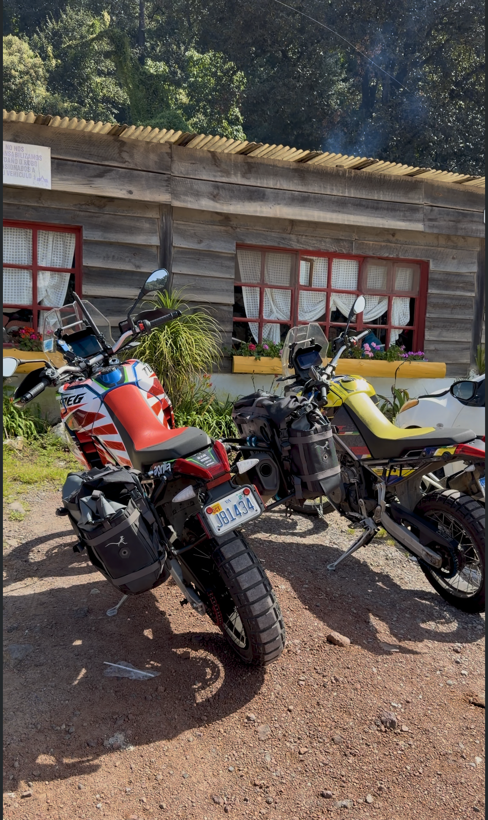 Two motorcycles parked outside a rustic wooden house with red-framed windows, surrounded by greenery and flower boxes.