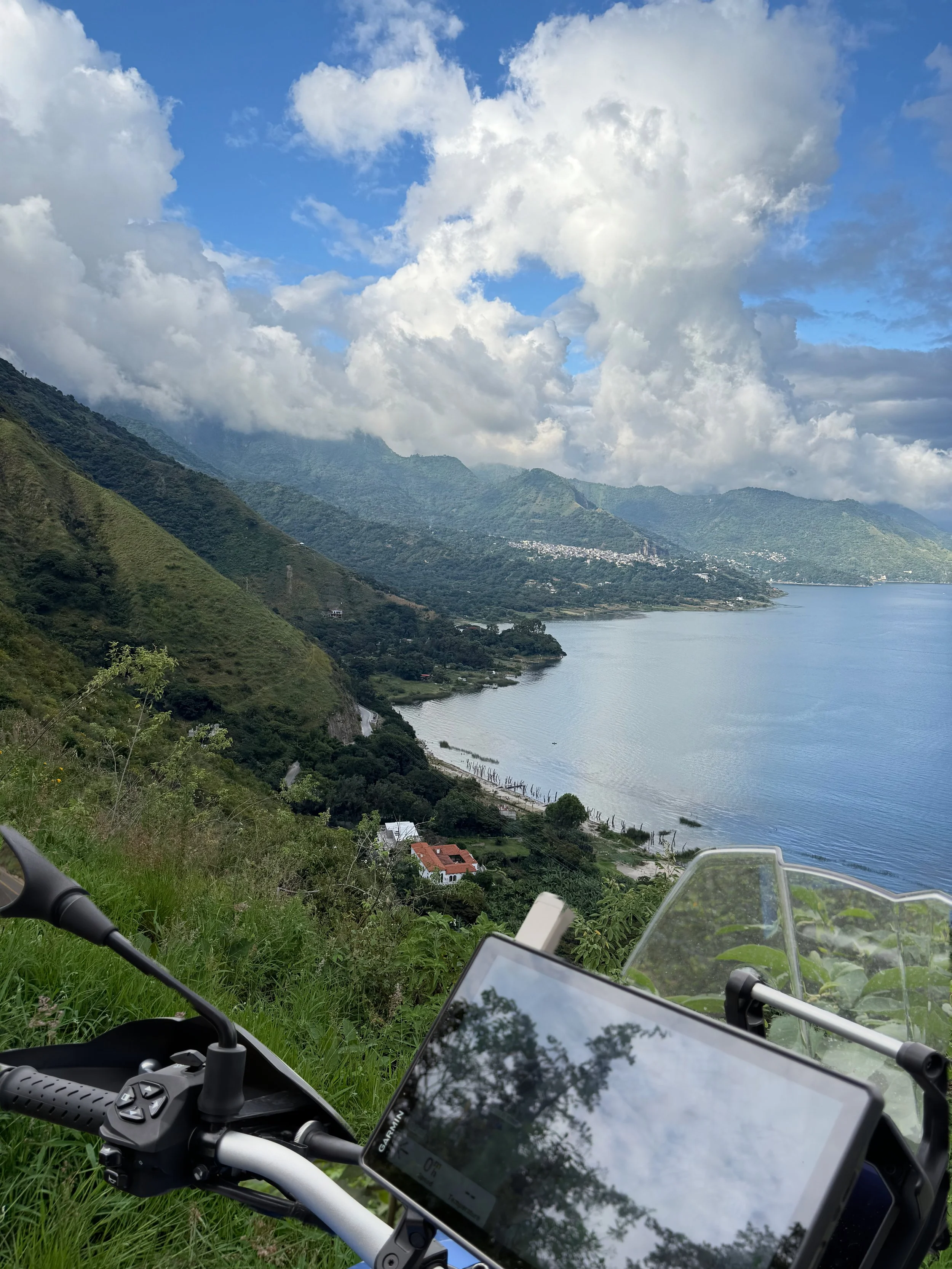 View from a motorcycle or bike along a hillside overlooking a lake with mountains and cloudy sky in the background. Part of the motorcycle and a GPS device are visible in the foreground.