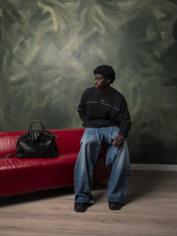 Seated Vanity Apparel editorial portrait in a black top and blue jeans against a green painted backdrop