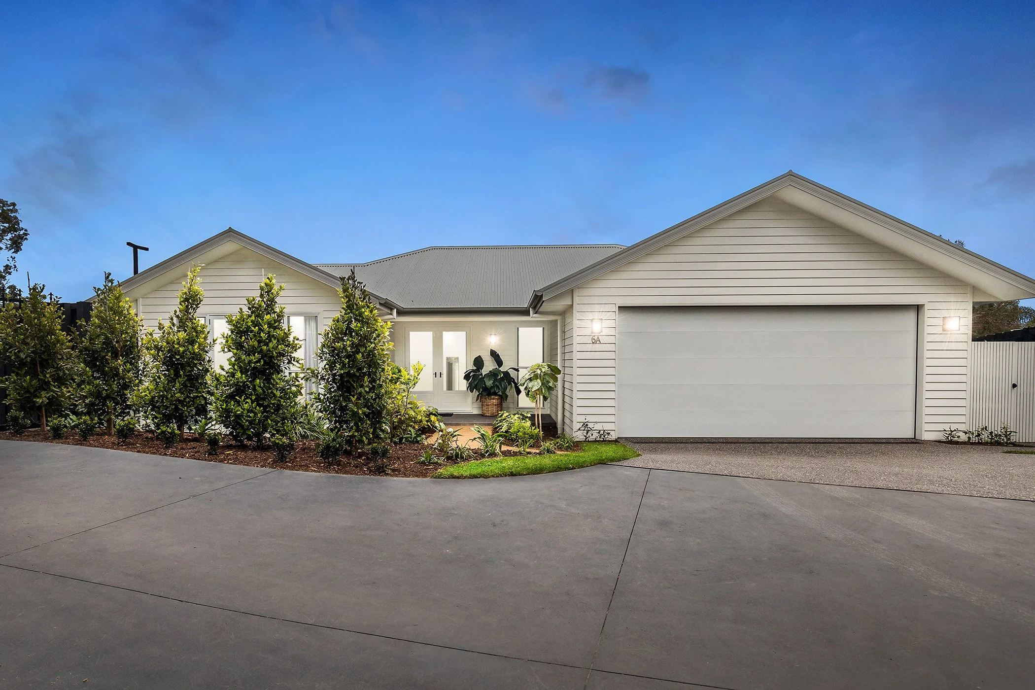Modern house with white siding, a garage door, front door, surrounded by greenery, under a clear evening sky.