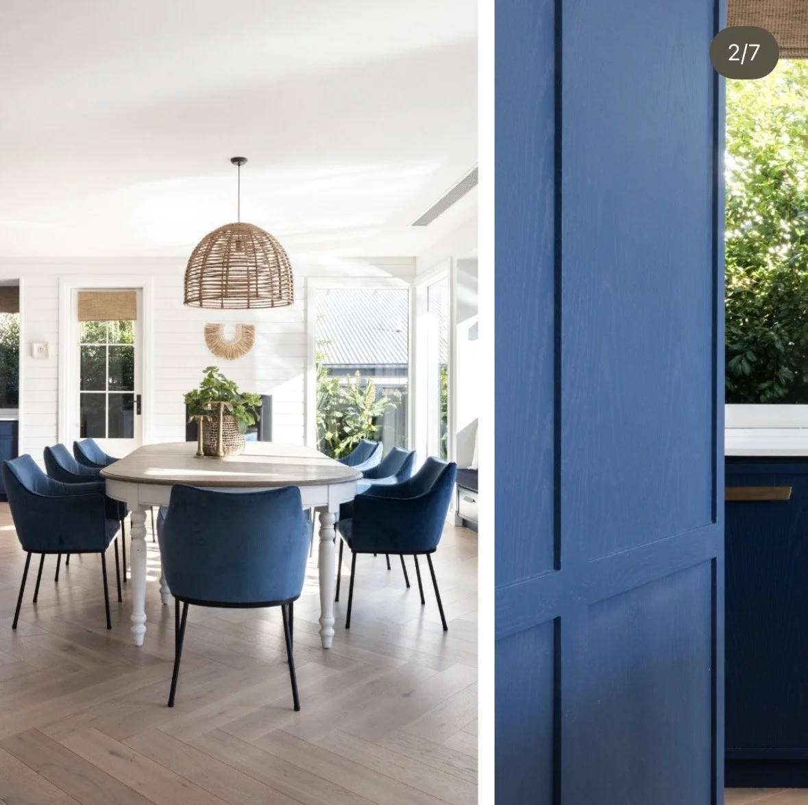Bright dining room with white walls, a large white oval table surrounded by dark blue chairs, a wicker lampshade hanging from the ceiling, a potted plant on the table, and large windows with greenery outside.
