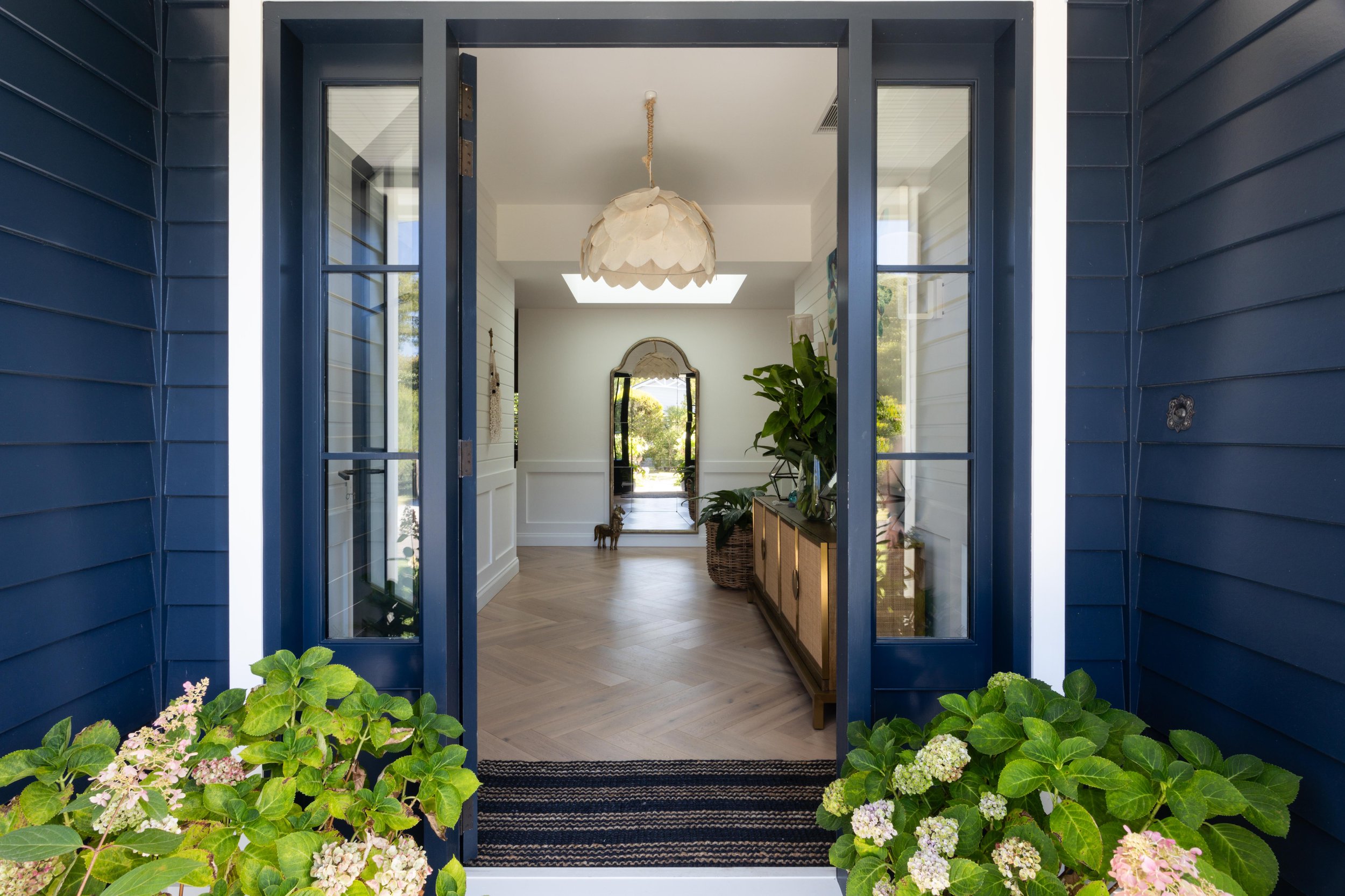 Entrance view from outside the house through a glass door with blue framing, leading into a bright, modern foyer with light wood flooring, a large potted plant, a gold-framed mirror, and a unique white pendant light. Green hydrangea flowers are visible at the bottom of the image.