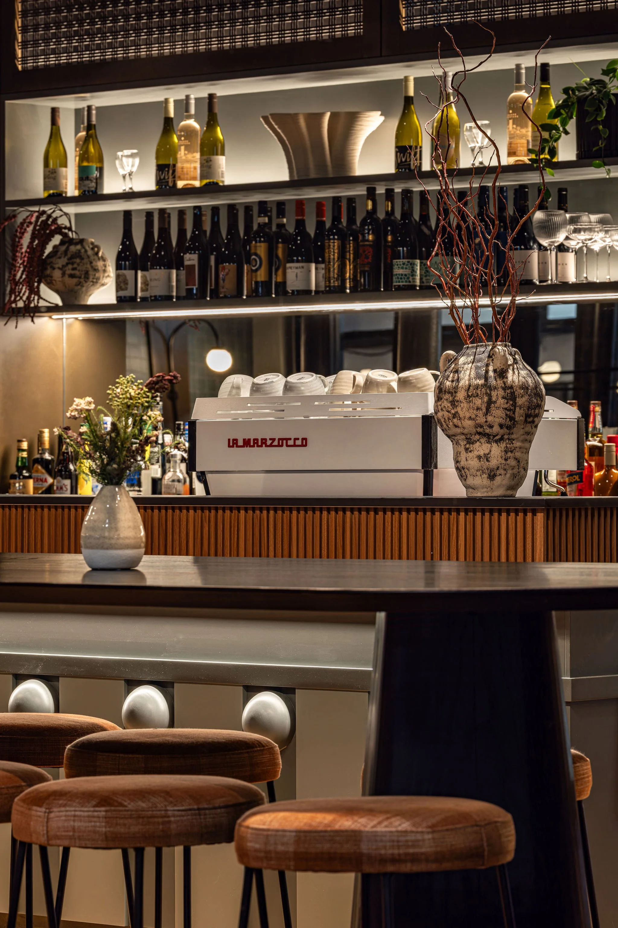 A bar interior with shelves of wine bottles, decorative vases, flowers, and bar stools in warm lighting.
