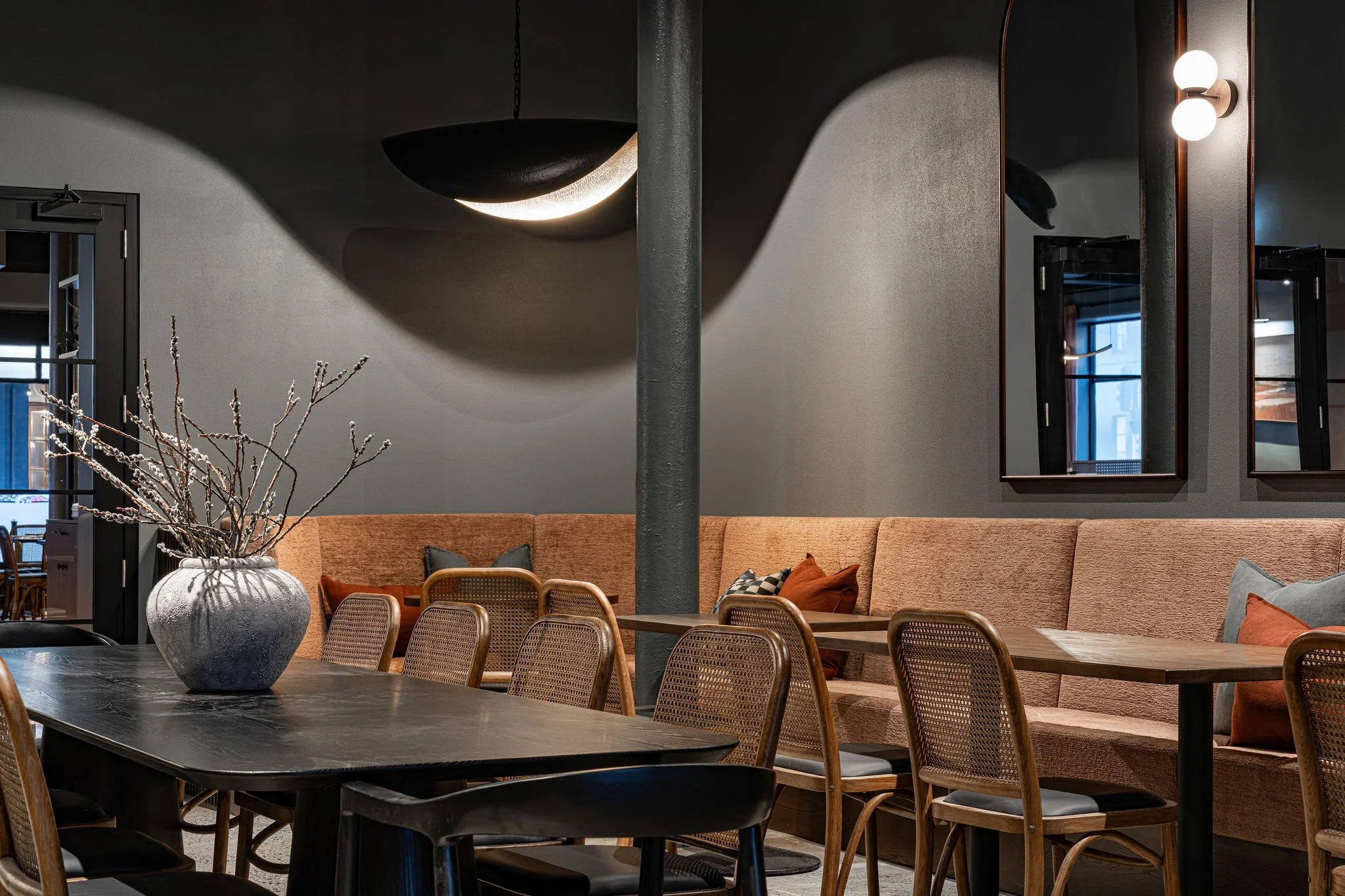 Interior of a modern dining space with a large black dining table, a vase with bare branches, and a light-colored upholstered banquette with pillows. There are wooden chairs, mirrors, and wall-mounted lights on the dark walls.