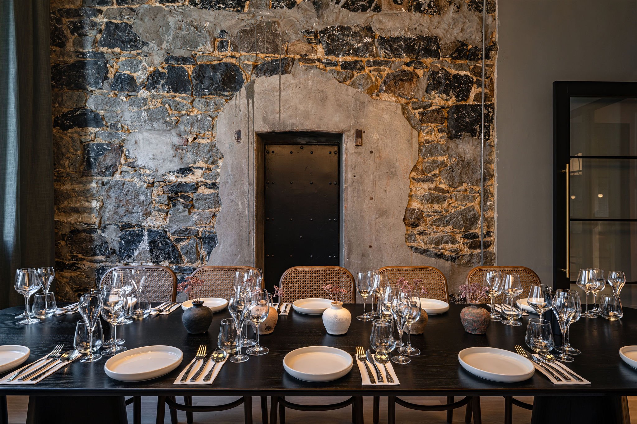 A dining room table set with white plates, wine glasses, and silverware. The table is in front of a rustic stone wall with an arched recess and a black door. The ambiance suggests a modern, stylish restaurant or event space.