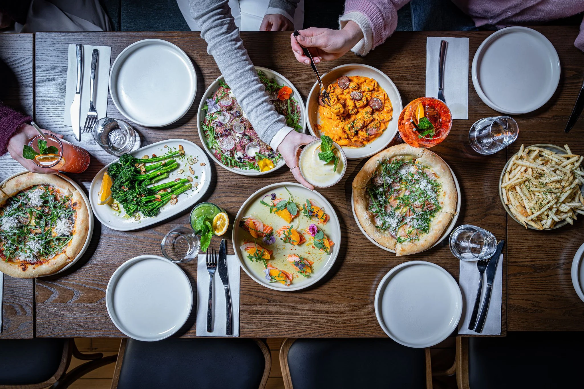 Top-down view of a table with various dishes, including two pizzettas, salads, pasta, and drinks, with empty plates and silverware set for guests.