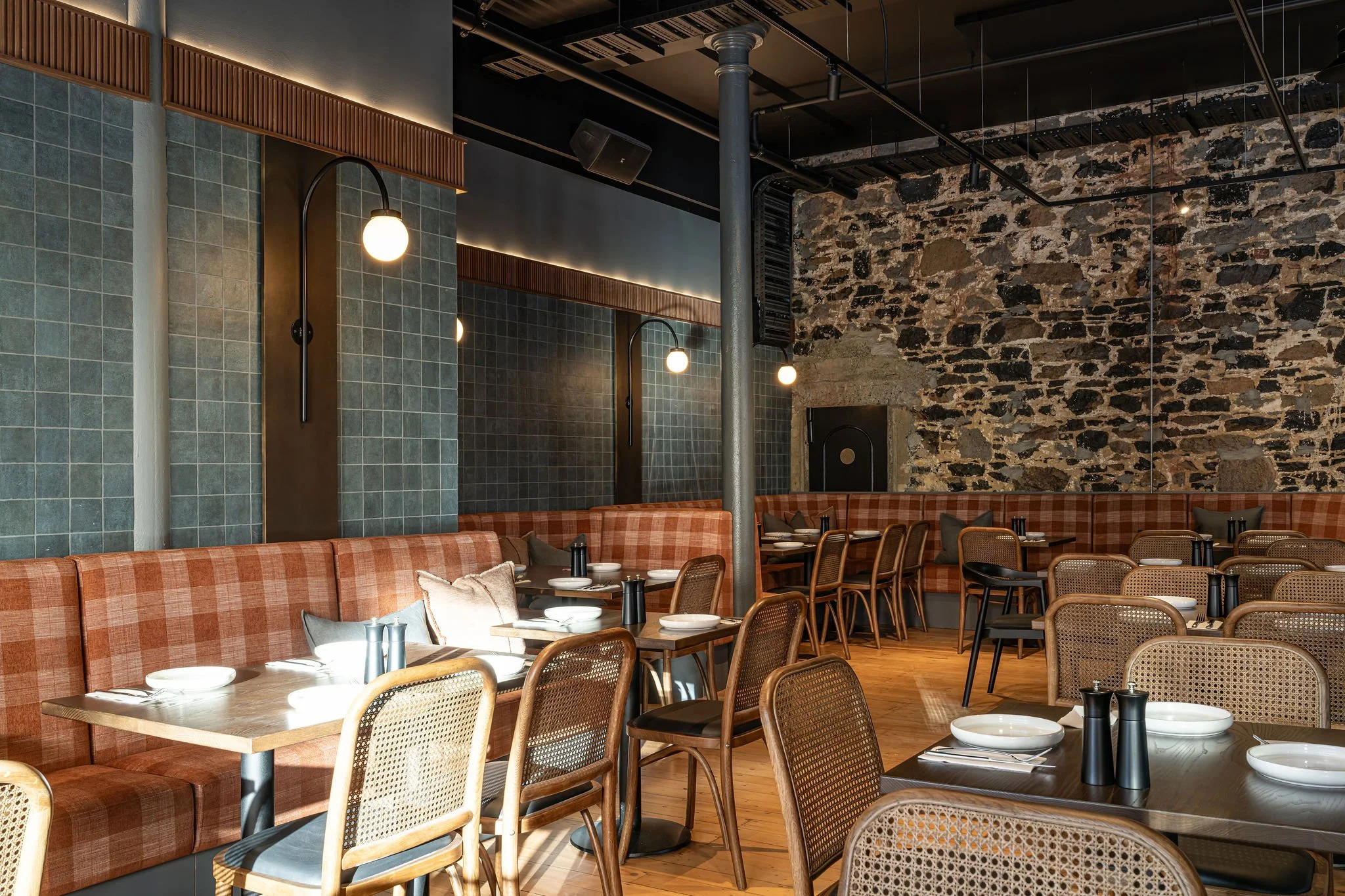 Interior of a modern restaurant with wooden tables, some set with plates, cups, and black condiment shakers, surrounded by chairs with woven backs. The walls feature green tiled and exposed stone sections, with wall sconces providing lighting. The ce