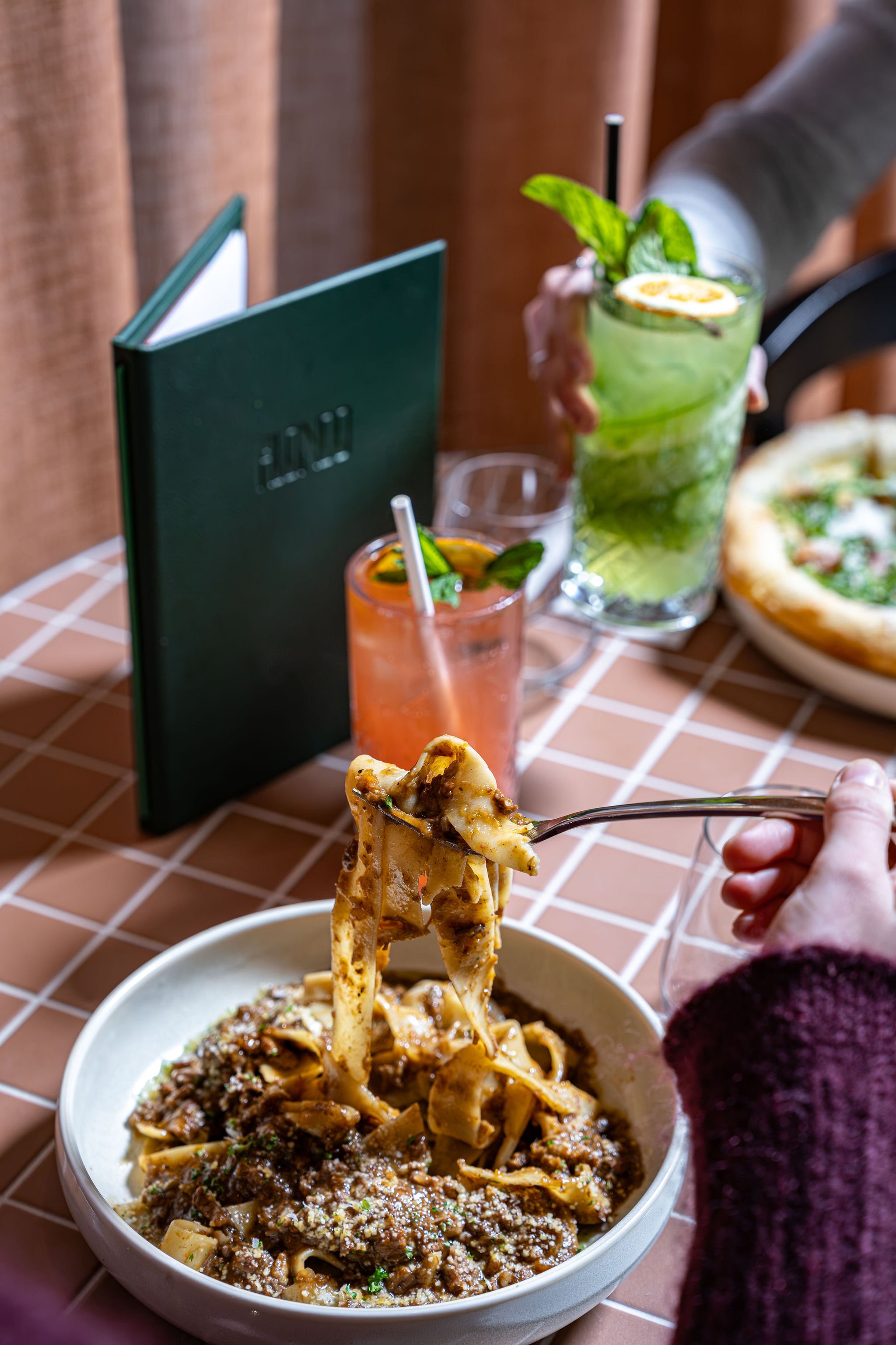 A person is holding a fork over a bowl of pappardelle with beef meat sauce. On the table, there are two drinks, one green and one orange, with garnishes. A pizzetta is partially visible in the background.