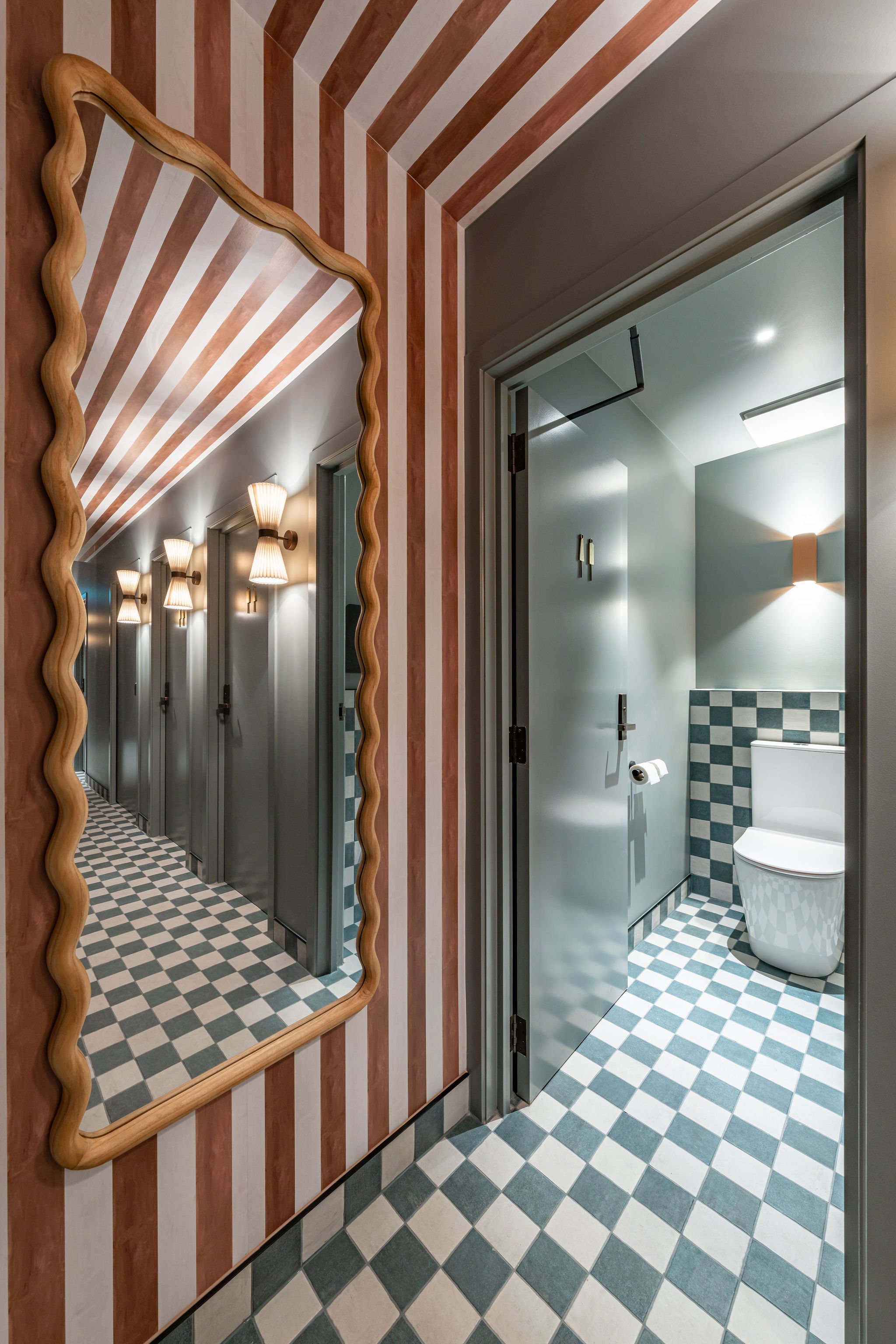 A bathroom with a checkered green and white floor, a toilet, and a mirror on the wall with a wavy wooden frame. The mirror reflects a hallway with striped wallpaper, wall-mounted lights, and having multiple doors.