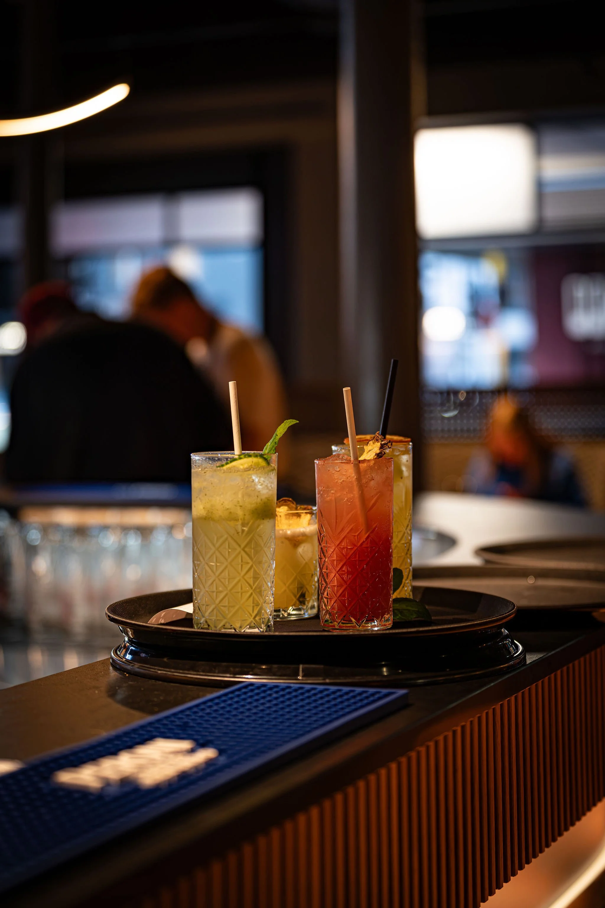 Three colorful cocktails with straws and garnishes are placed on a black tray on a bar counter, with a blurred background of people and bar interior.