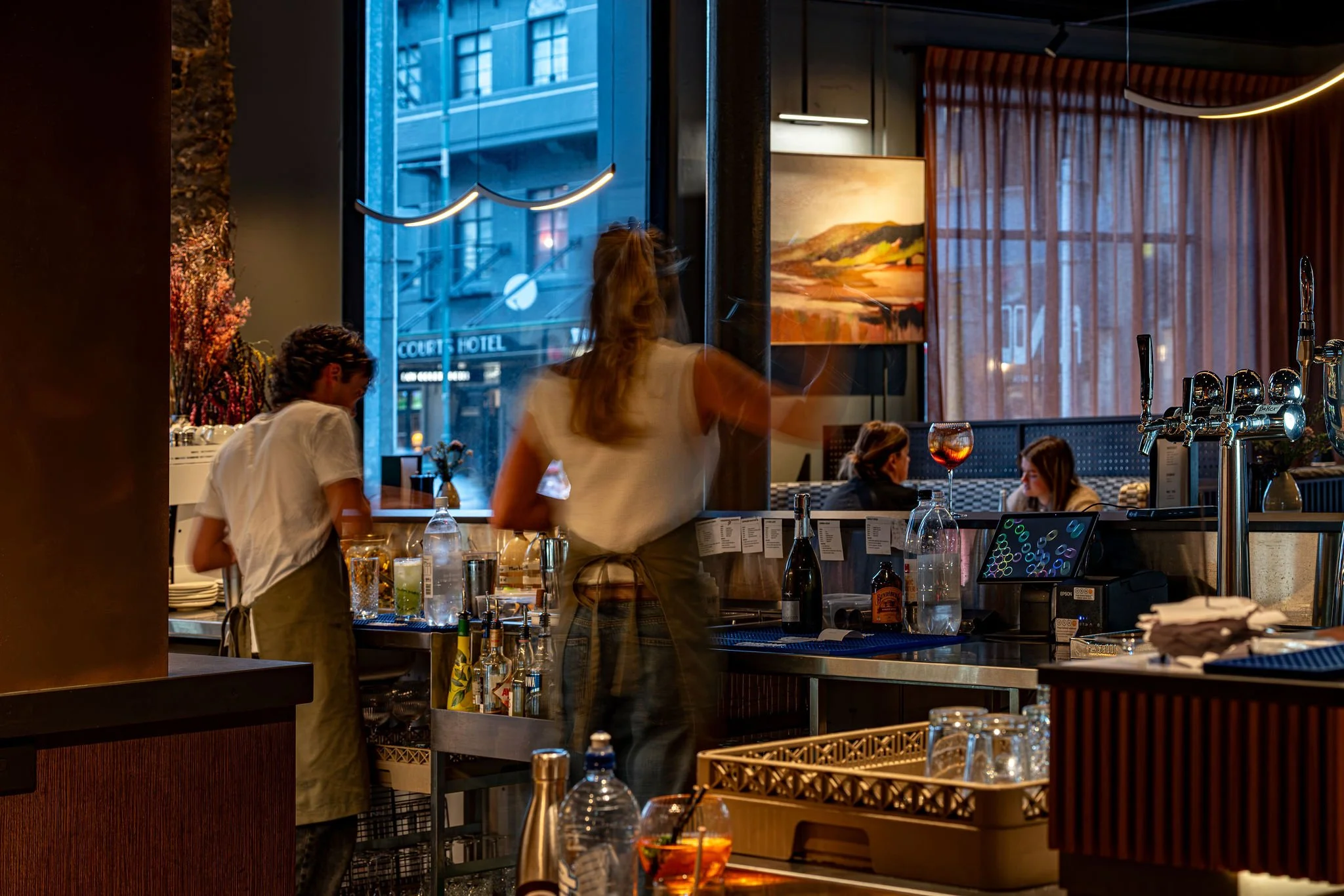 Interior of a restaurant or bar with large windows showing city buildings outside. Several people are inside, including a woman with long hair and a man with curly hair. There are glasses, bottles, and bar equipment on the counter, and artwork hangs on the wall. Warm lighting and sheer curtains add to the cozy atmosphere.