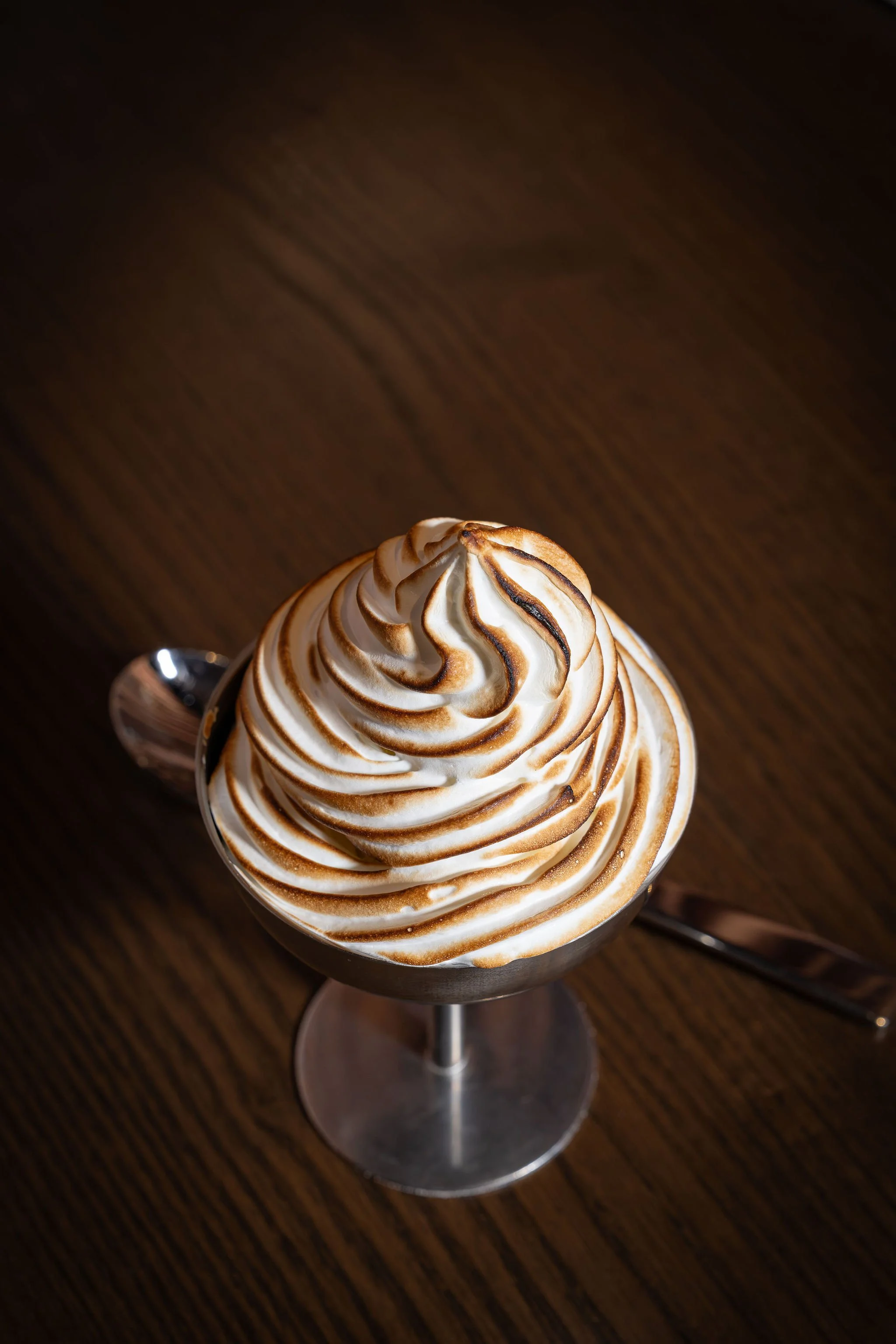 A glass dessert cup filled with whipped cream topped with toasted marshmallow meringue with unburned brown streaks, garnished with a toasted marshmallow on top, placed on a wooden table.