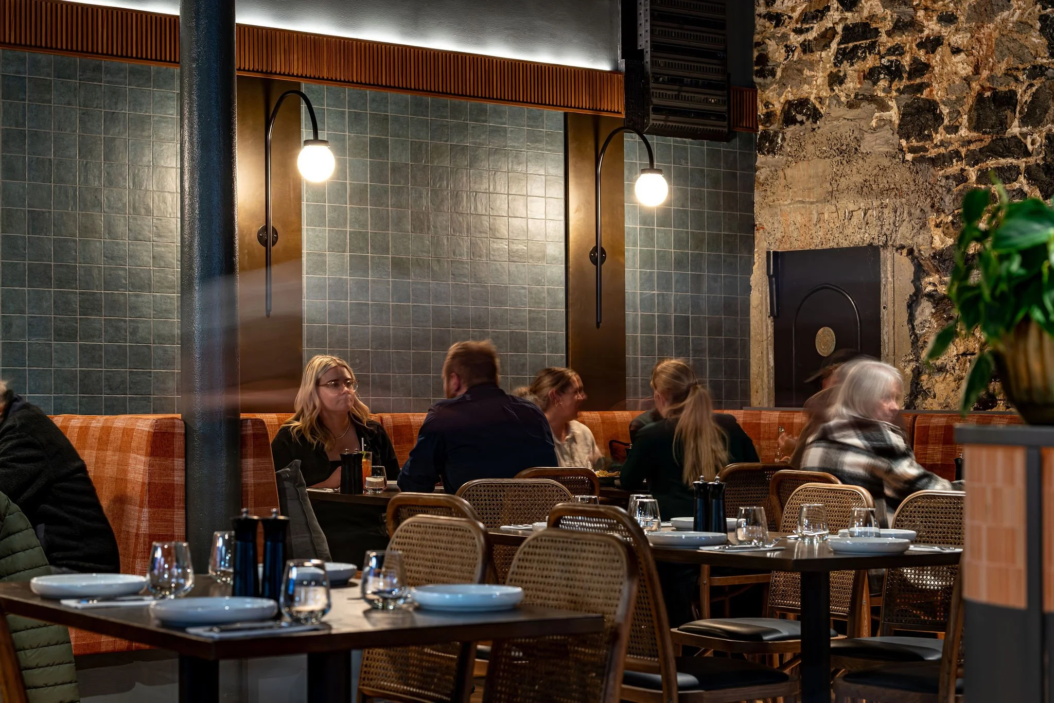 Interior of a restaurant with people sitting at tables, some in conversation. The walls are decorated with dark green tiles and a stone wall. There are black wall lights, and wooden accents are visible.