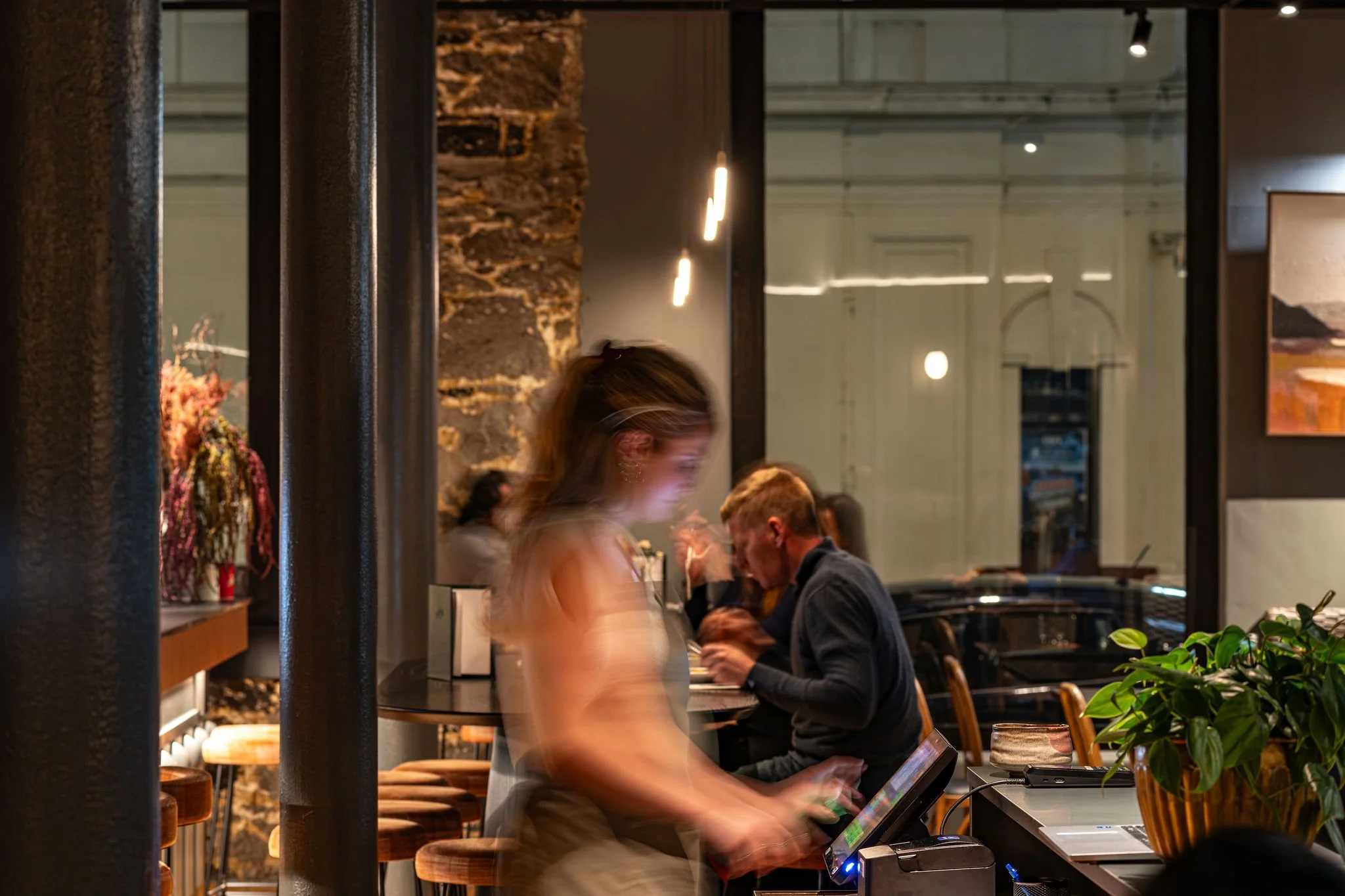 People dining in a restaurant with warm lighting, exposed brick walls, and greenery. A woman appears blurry in motion, sitting near a man reading from a menu.