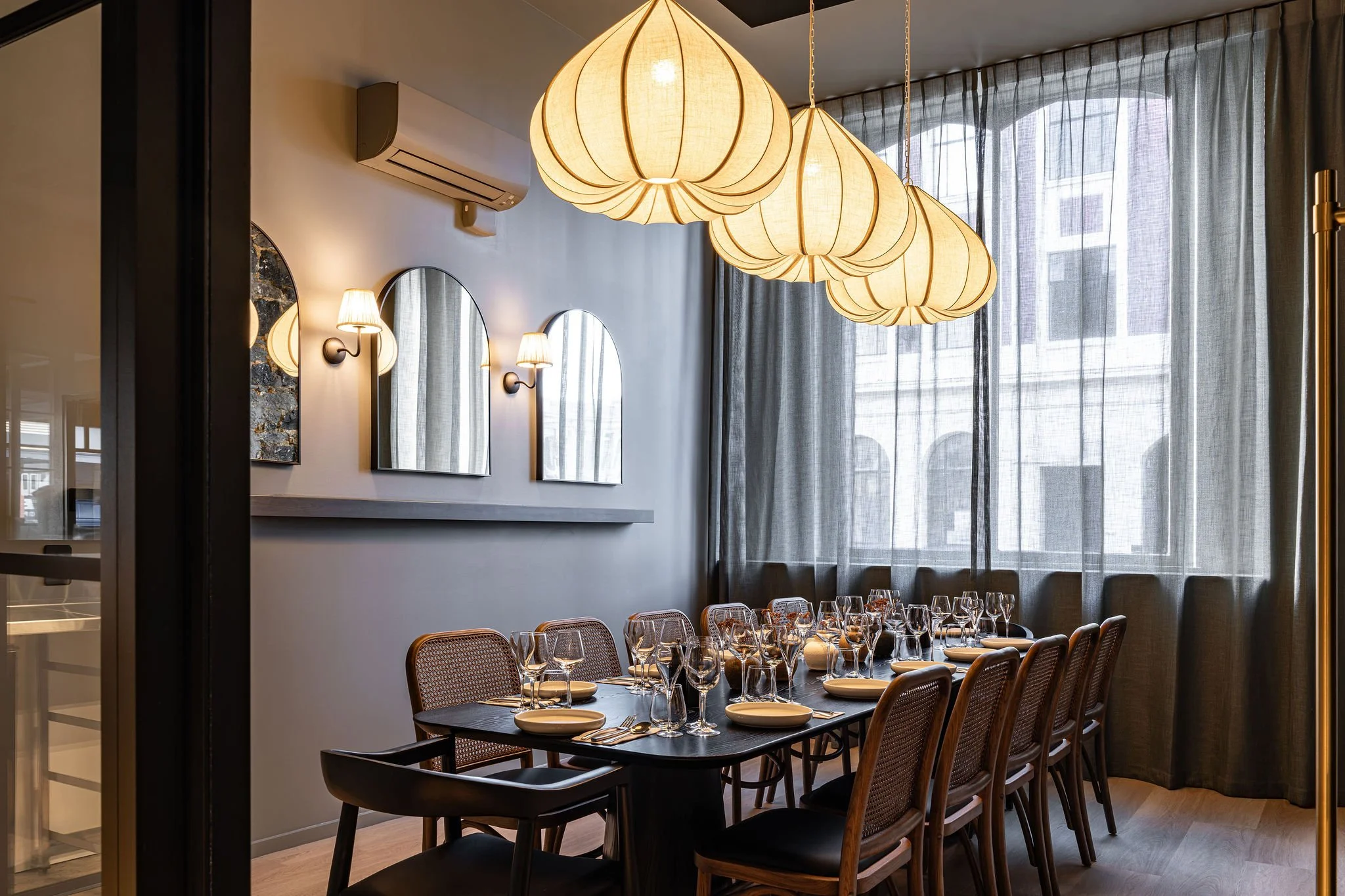 Elegant dining room with hanging pendant lights, wall sconces, a long table set with wine glasses, plates, and silverware, and large windows with sheer curtains.