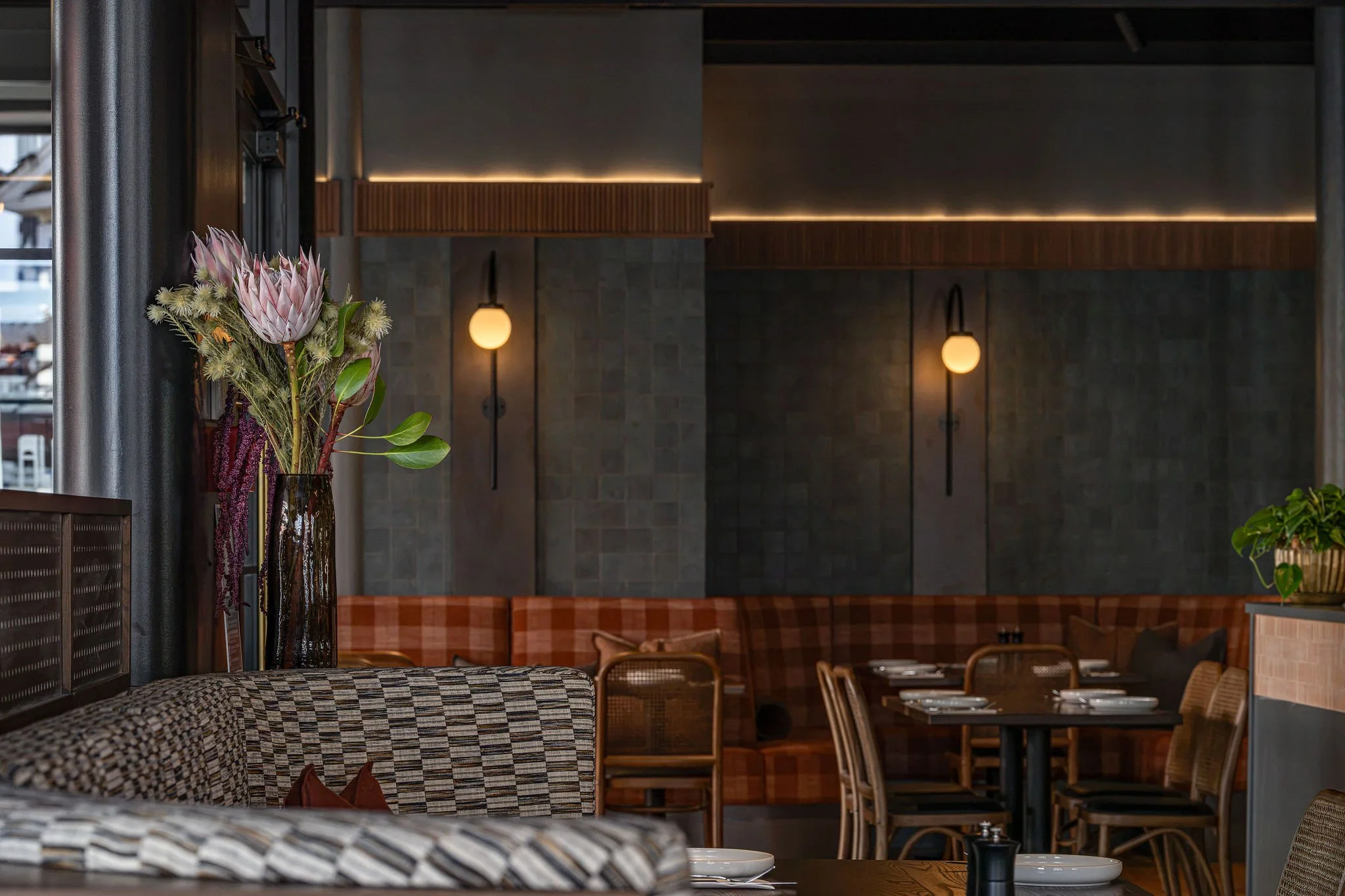 Interior of a restaurant with a large flower arrangement on a table, wooden chairs, and modern lighting fixtures on the walls.