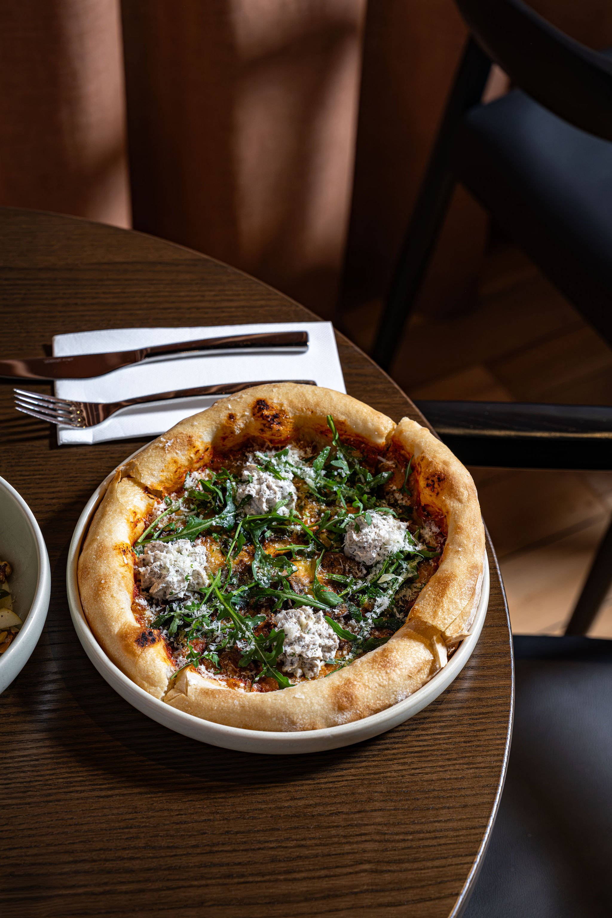 A plate of pizzetta topped with arugula and dollops of cheese on a wooden table, with a knife and fork on a white napkin beside it, and a dark chair in the background.