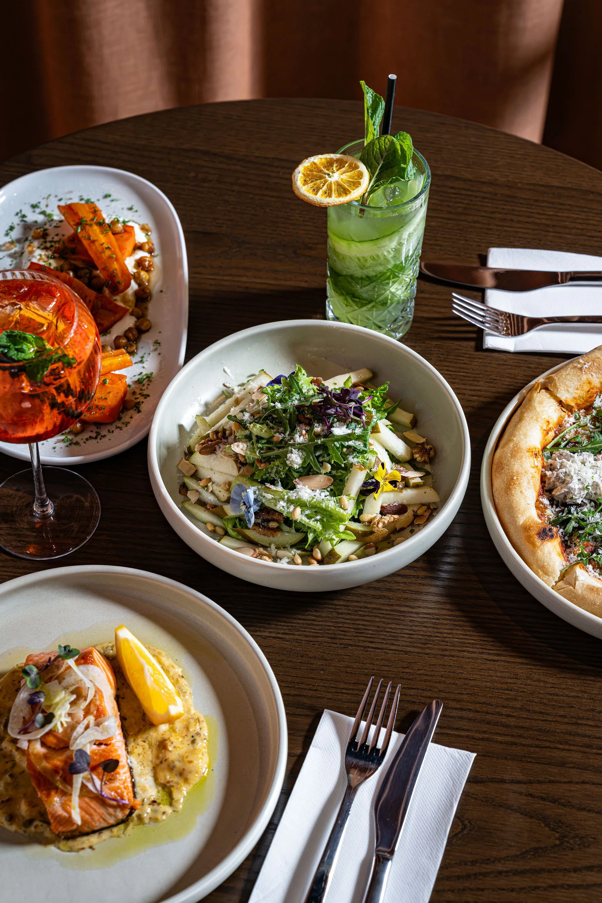 A close-up view of a dining table with various dishes including a salad, pizzetta, and another dish with salmon and lemon slices, along with a tall glass of green beverage garnished with lemon and mint, set on a wooden table with cutlery and napkins.