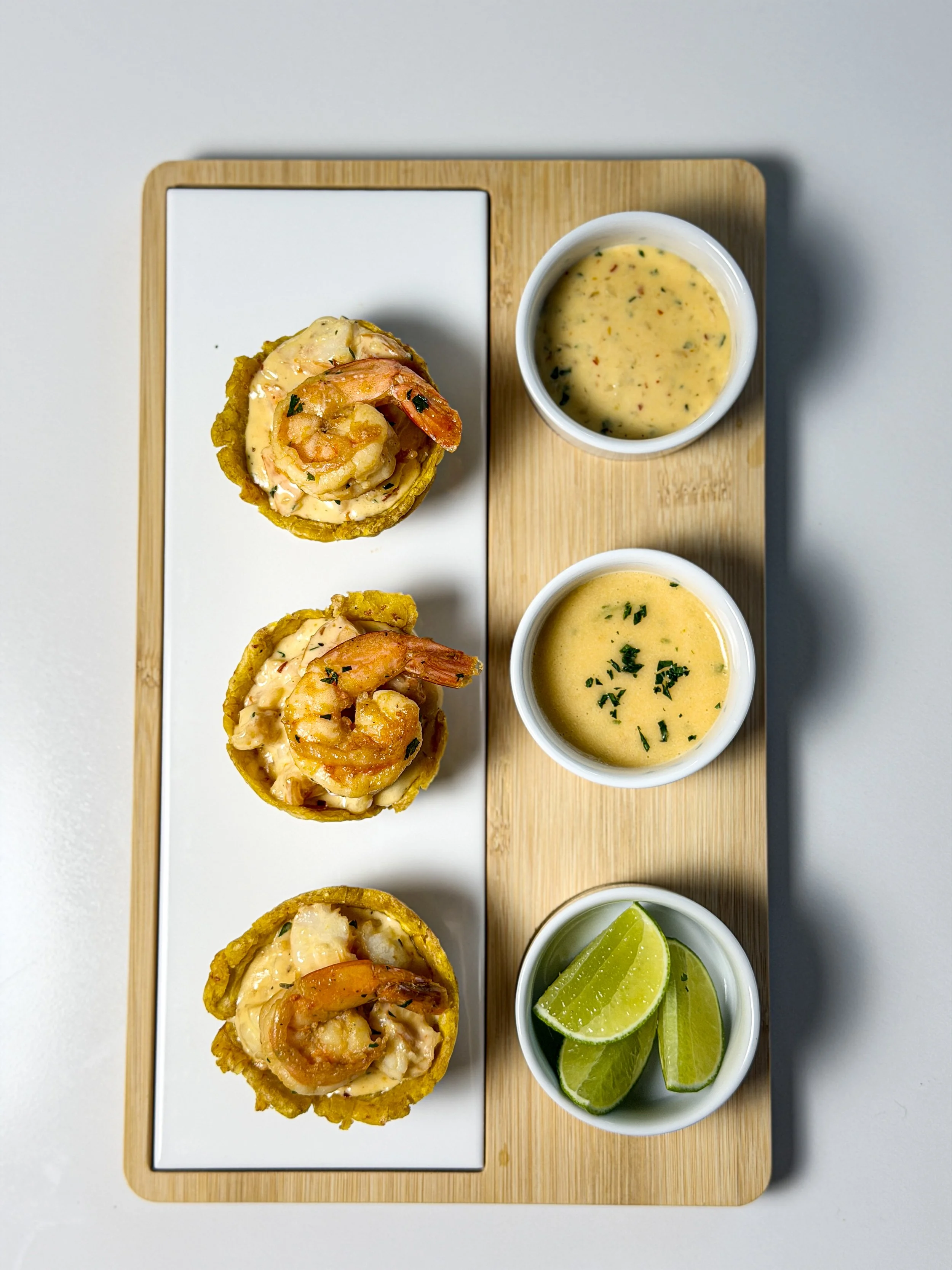 Three shrimp and seafood appetizers in cups with lime wedges, served with two bowls of dipping sauces on a wooden platter.