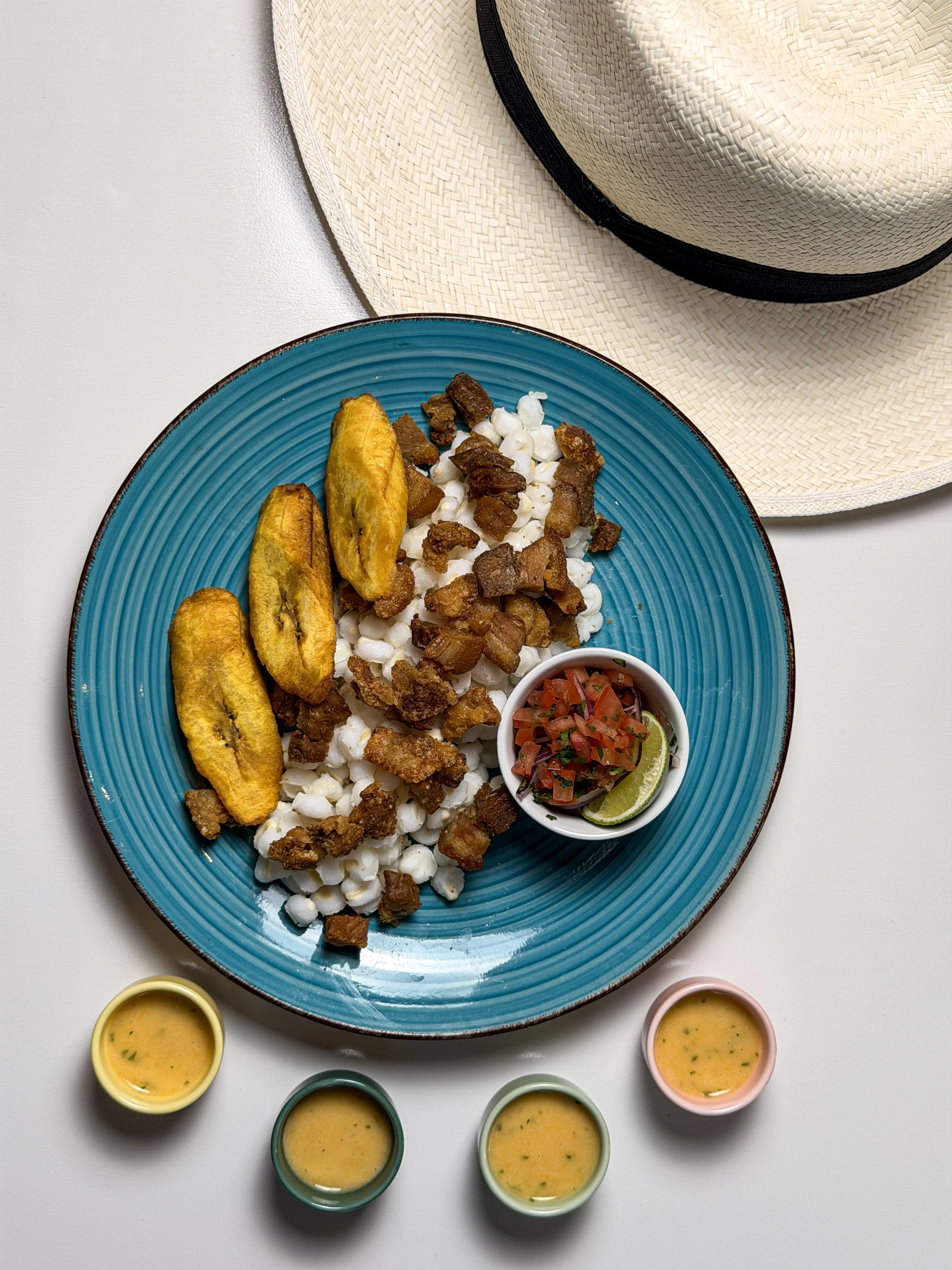 A blue plate containing fried plantains, crumbled cheese, and chopped pork, with a small bowl of salsa and lime, and four small cups of sauce or dressing nearby, on a white surface with a tan straw hat in the background.