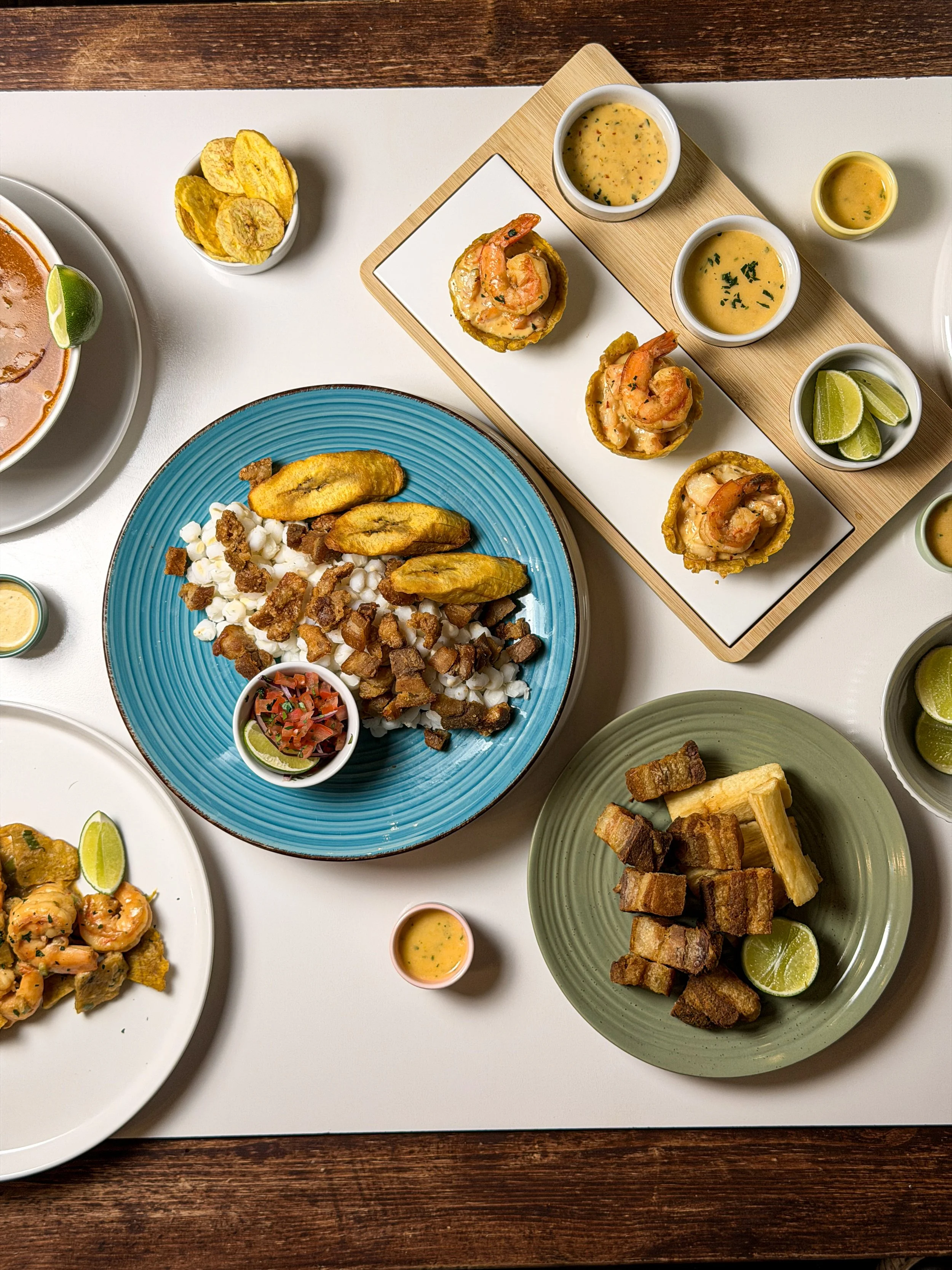 Assorted plates of Latin American dishes, including deep-fried plantains, pork cracklings, shrimp, and dipping sauces, on a white table.