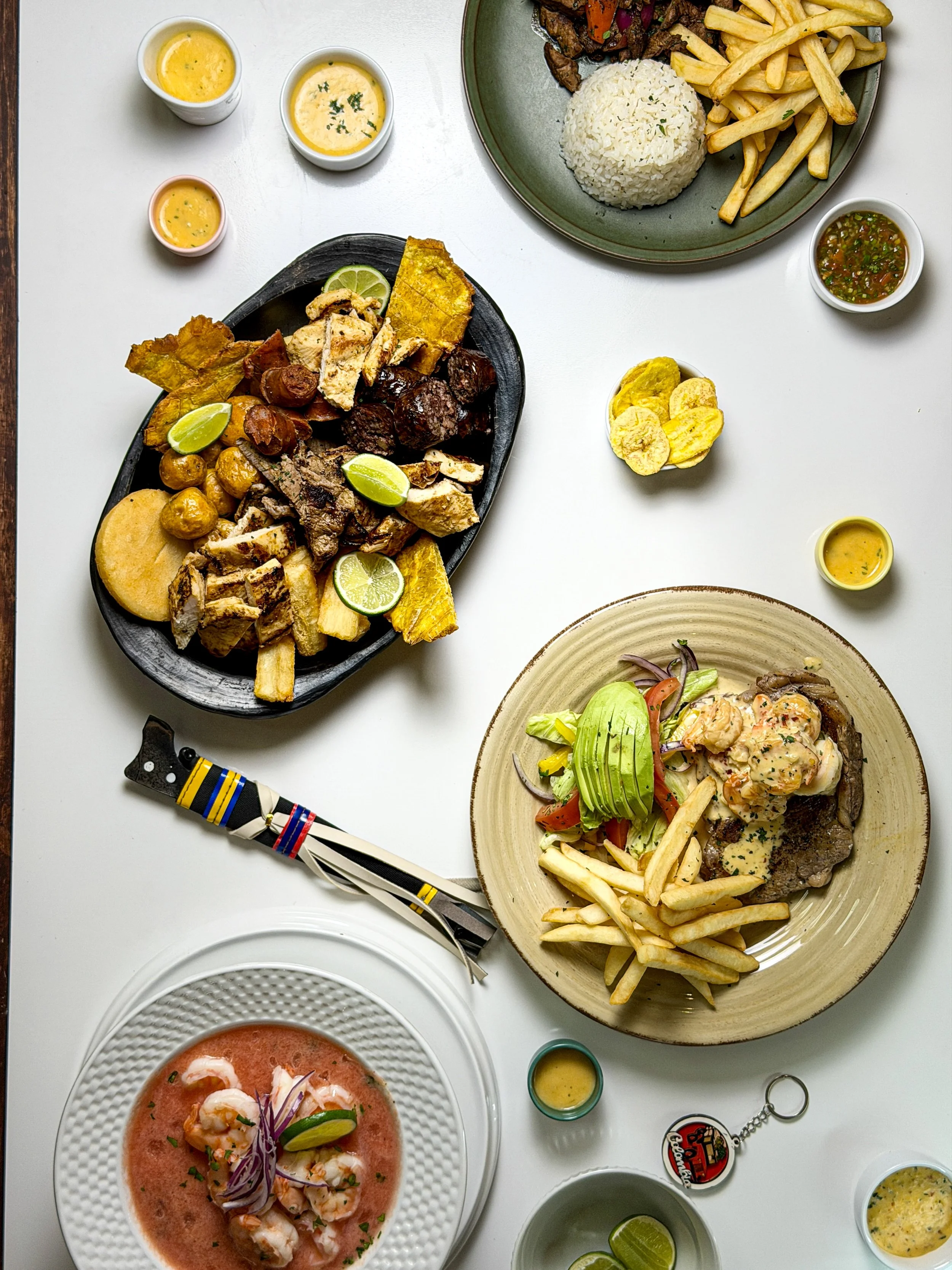 An overhead view of a table with various dishes including a seafood ceviche, a plate with steak, fries, and salad, a charcuterie board with meats and cheeses, bowls of soup, and small cups of sauce and lime wedges.