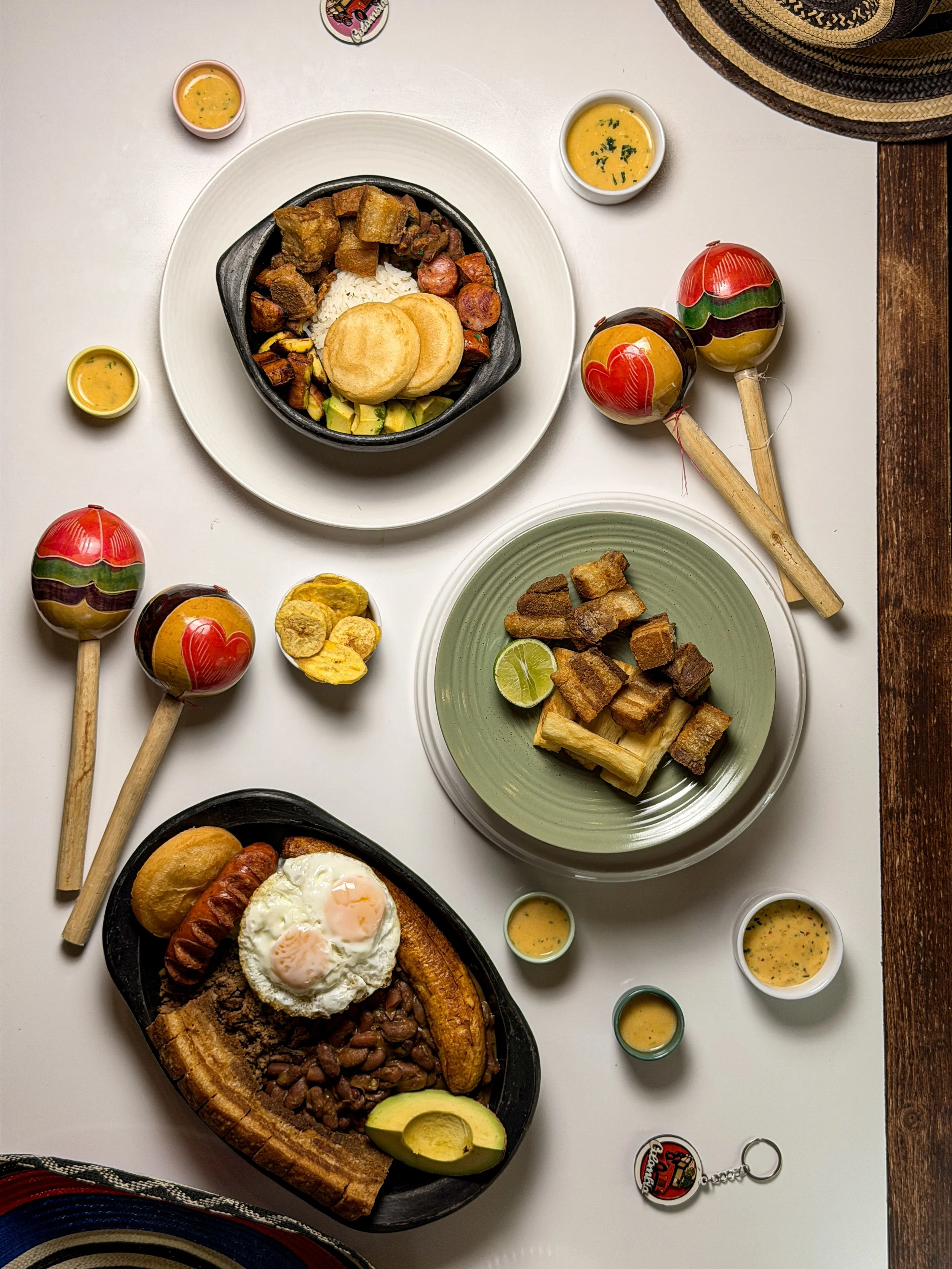 Overhead view of a variety of traditional Colombian foods on a white table, including bandeja paisa with rice, beans, fried eggs, chicharrón, and plantains, surrounded by small bowls of sauce, colorful Colombian hats, maracas, a keychain, and prepare