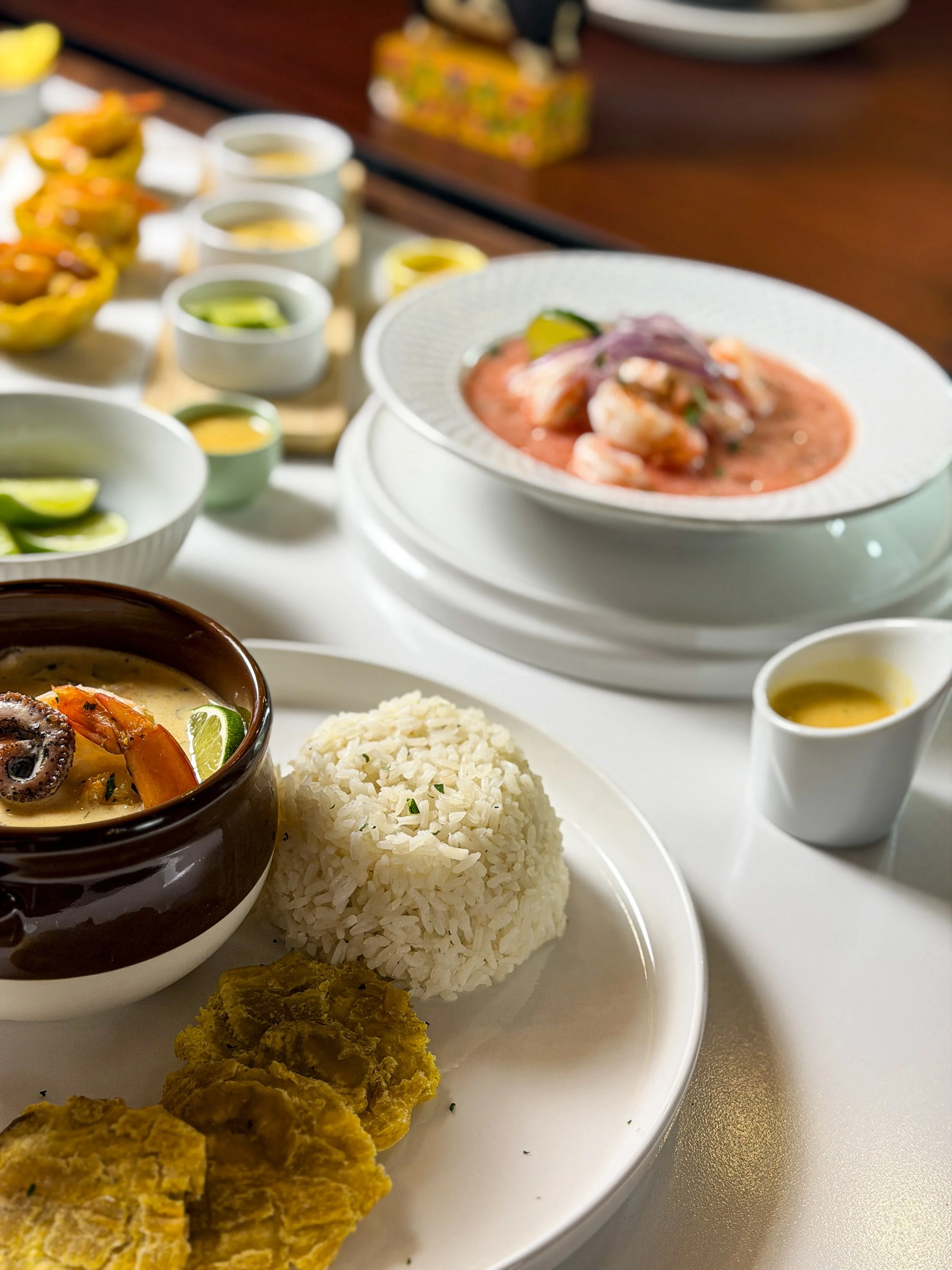 Assorted Latin American dishes on a table including rice, oxtail stew, fried plantains, and a shrimp ceviche, with small bowls of dipping sauces.