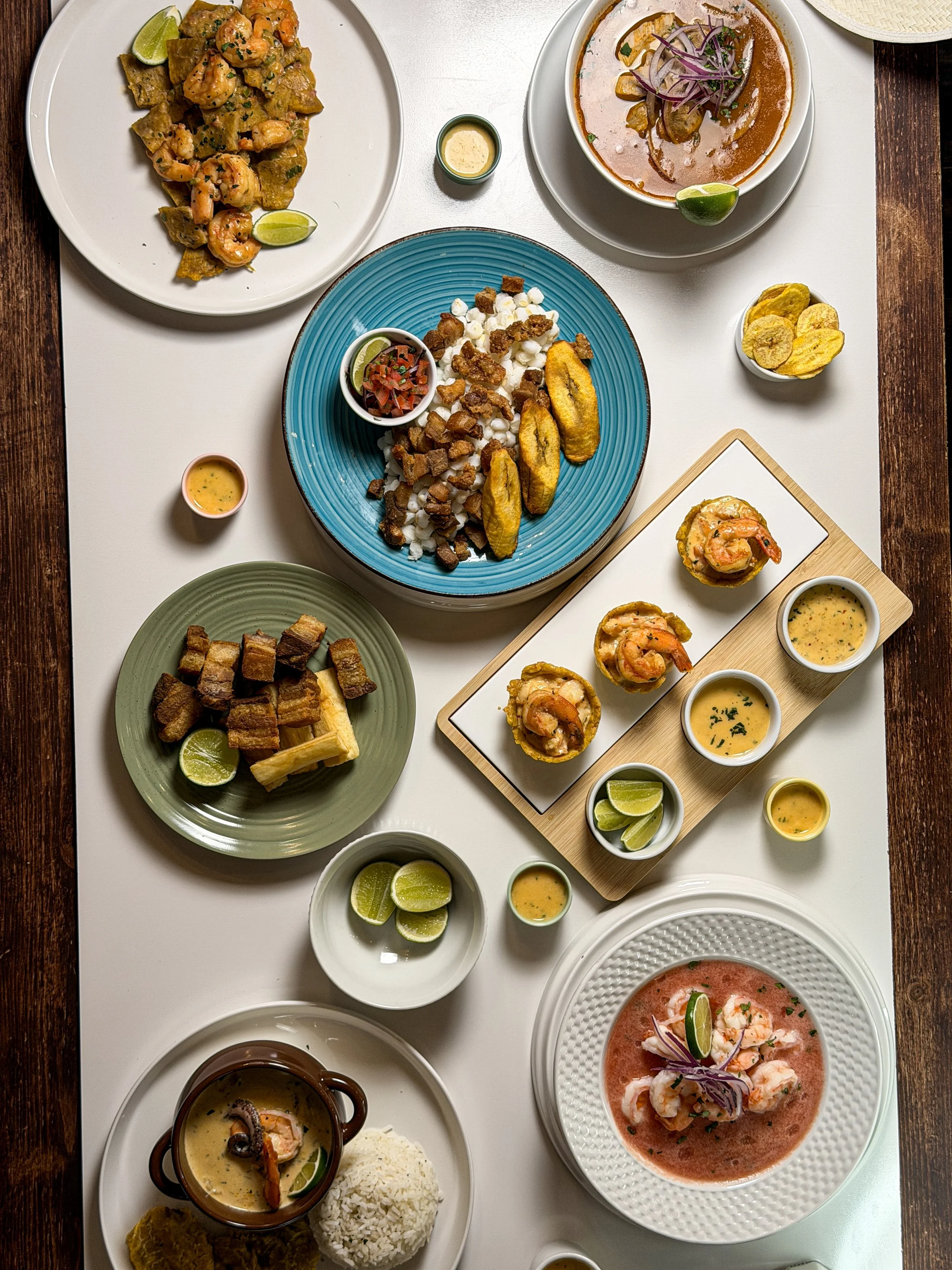 A variety of South American dishes on a white table, including shrimp, ceviche, grilled meats, soups, and various sides with lime wedges and dipping sauces.