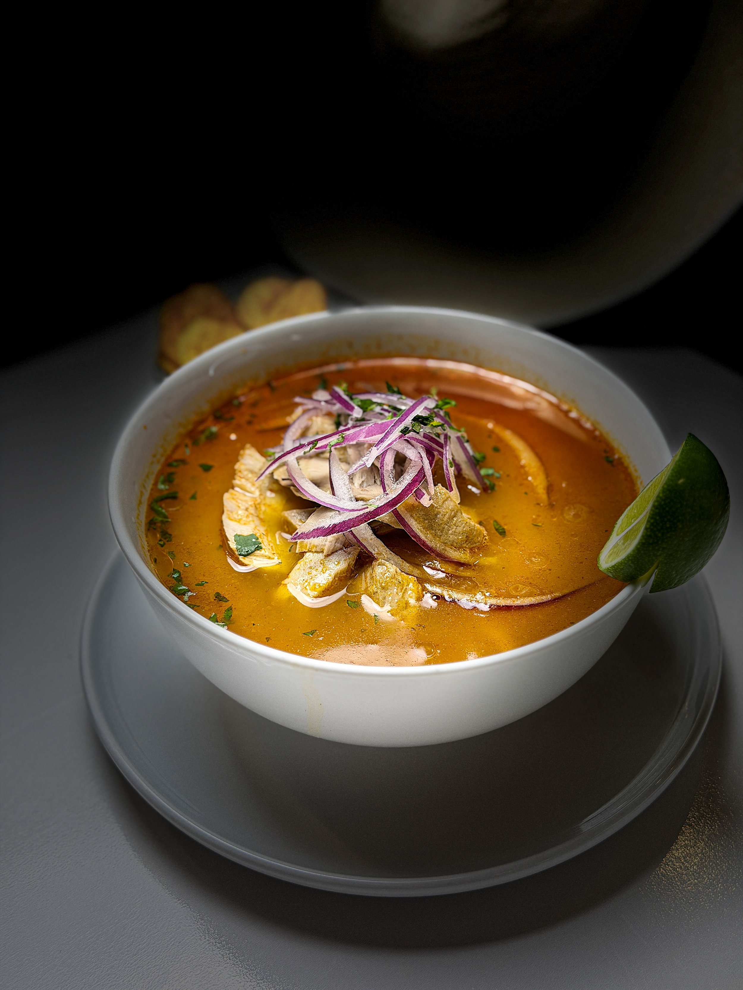 A bowl of creamy seafood soup garnished with sliced red onions, herbs, and a lime wedge, with a spoon and rolled chips in the background.
