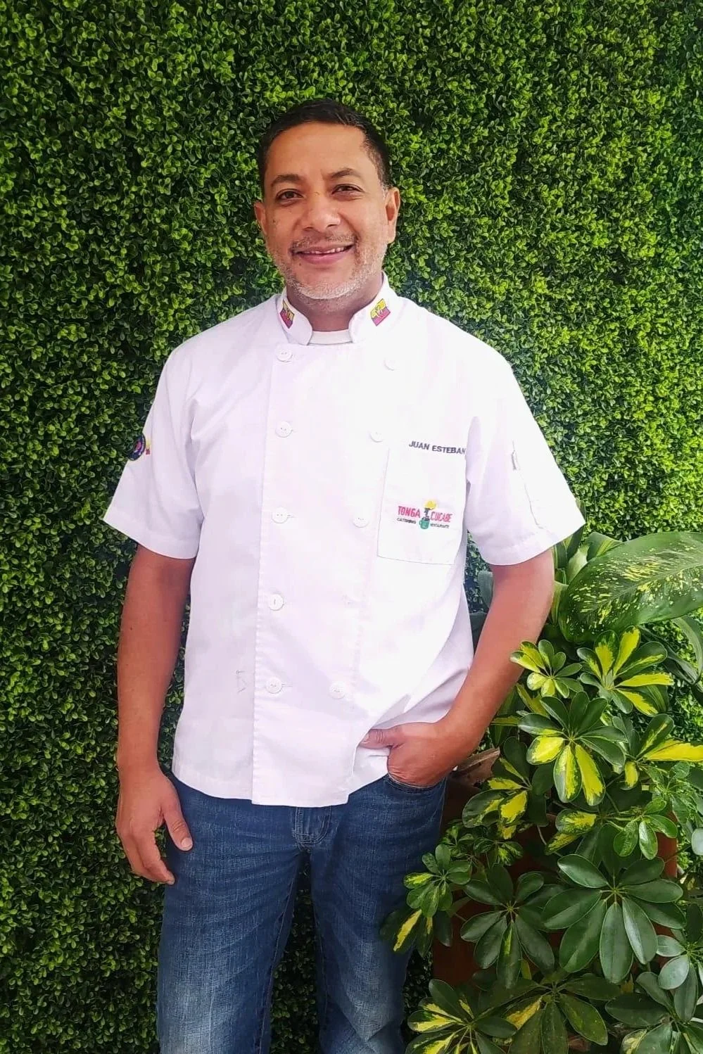A man in a white chef's jacket standing outdoors in front of a green leafy hedge and plants.