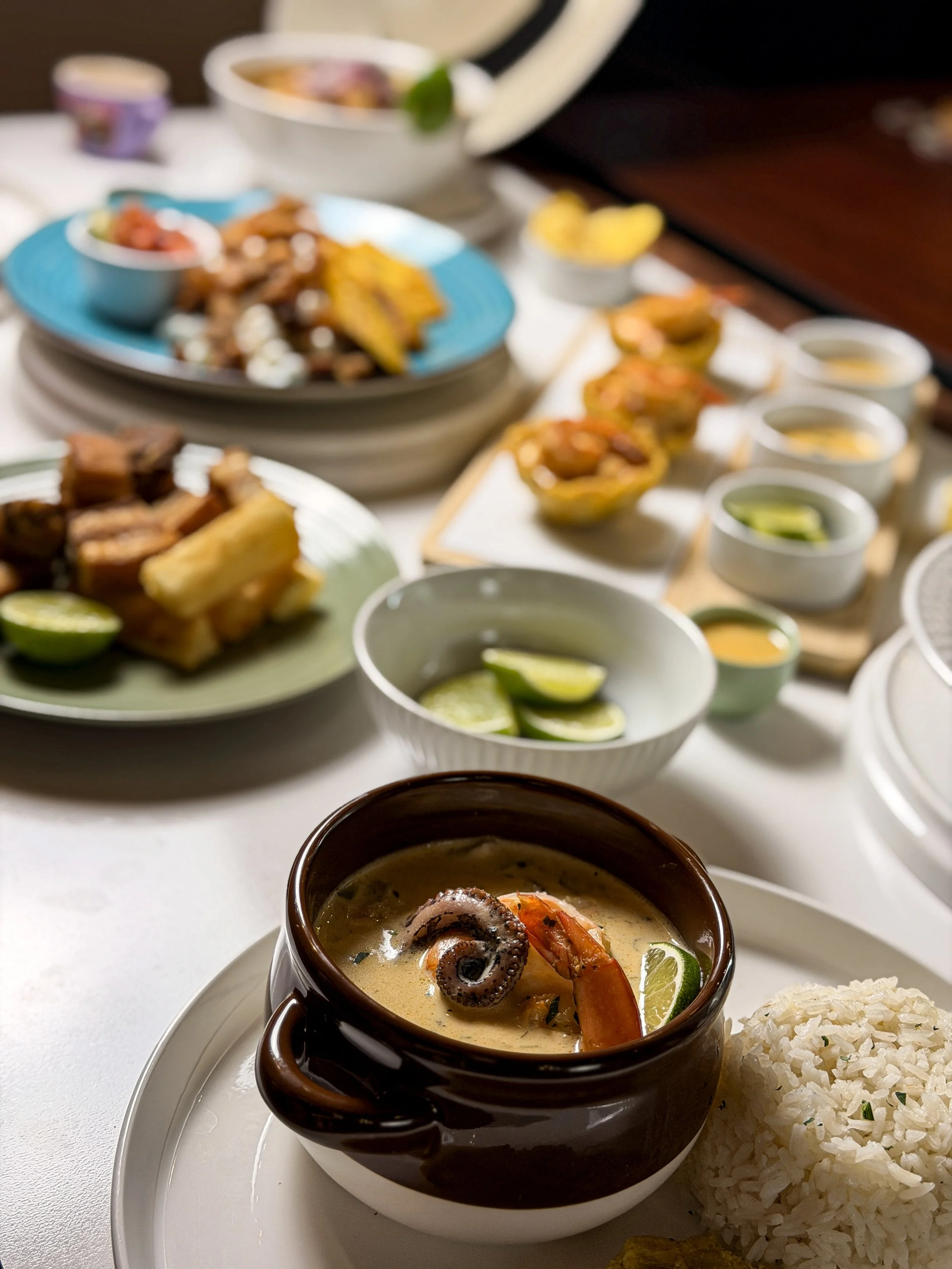 A table filled with Mexican dishes including a bowl of seafood soup with shrimp, octopus, and lime, a serving of white rice, small bowls of lime wedges, and various tacos and appetizers in the background.