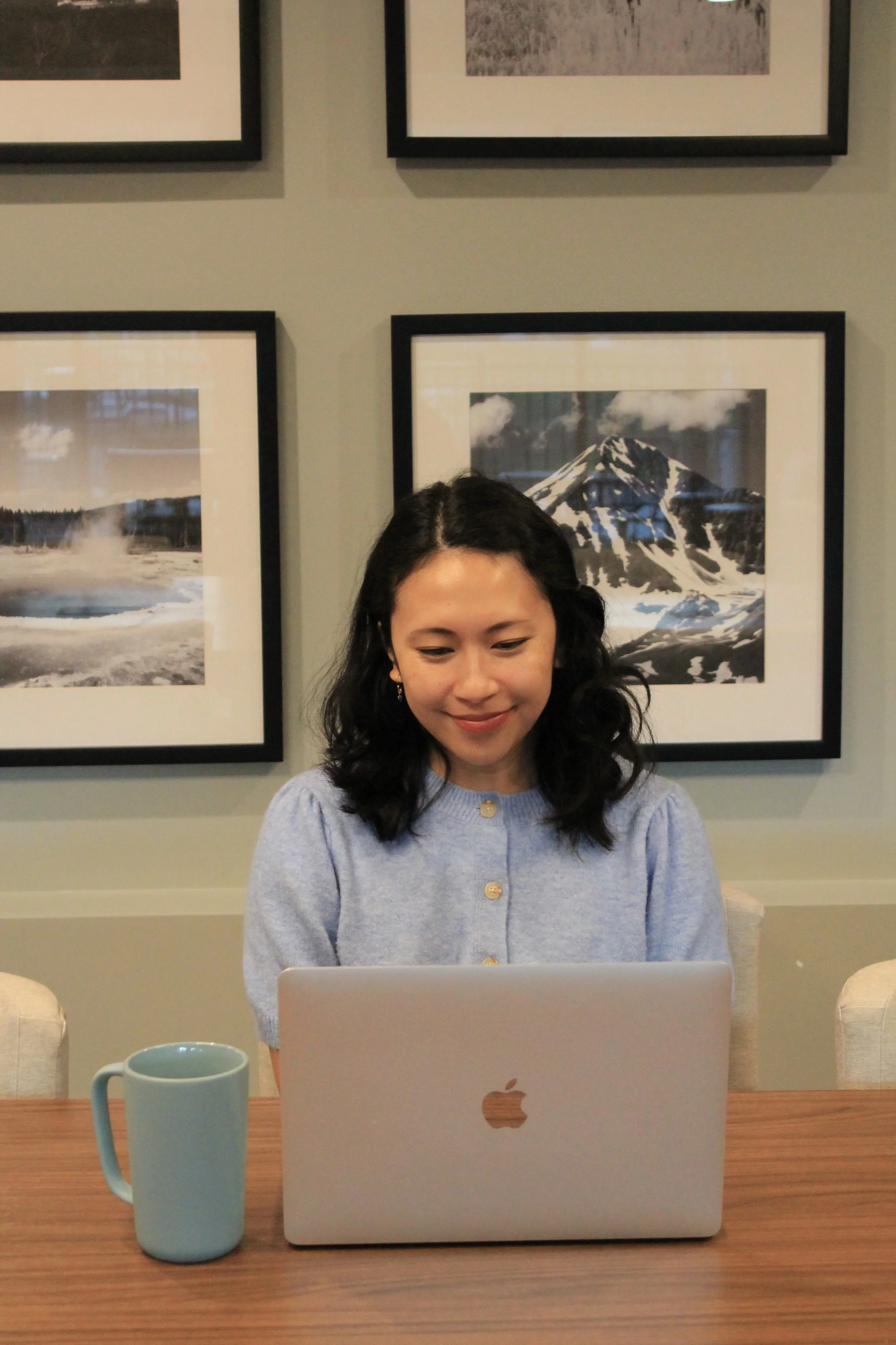 A young woman with dark, curly hair is sitting at a wooden table, working on a silver Apple MacBook laptop. She has a gentle smile and is wearing a light blue sweater with buttons. In front of her is a large, light blue mug. The background includes framed black and white photographs of nature scenes, featuring mountains and a geothermal area, on a beige wall.