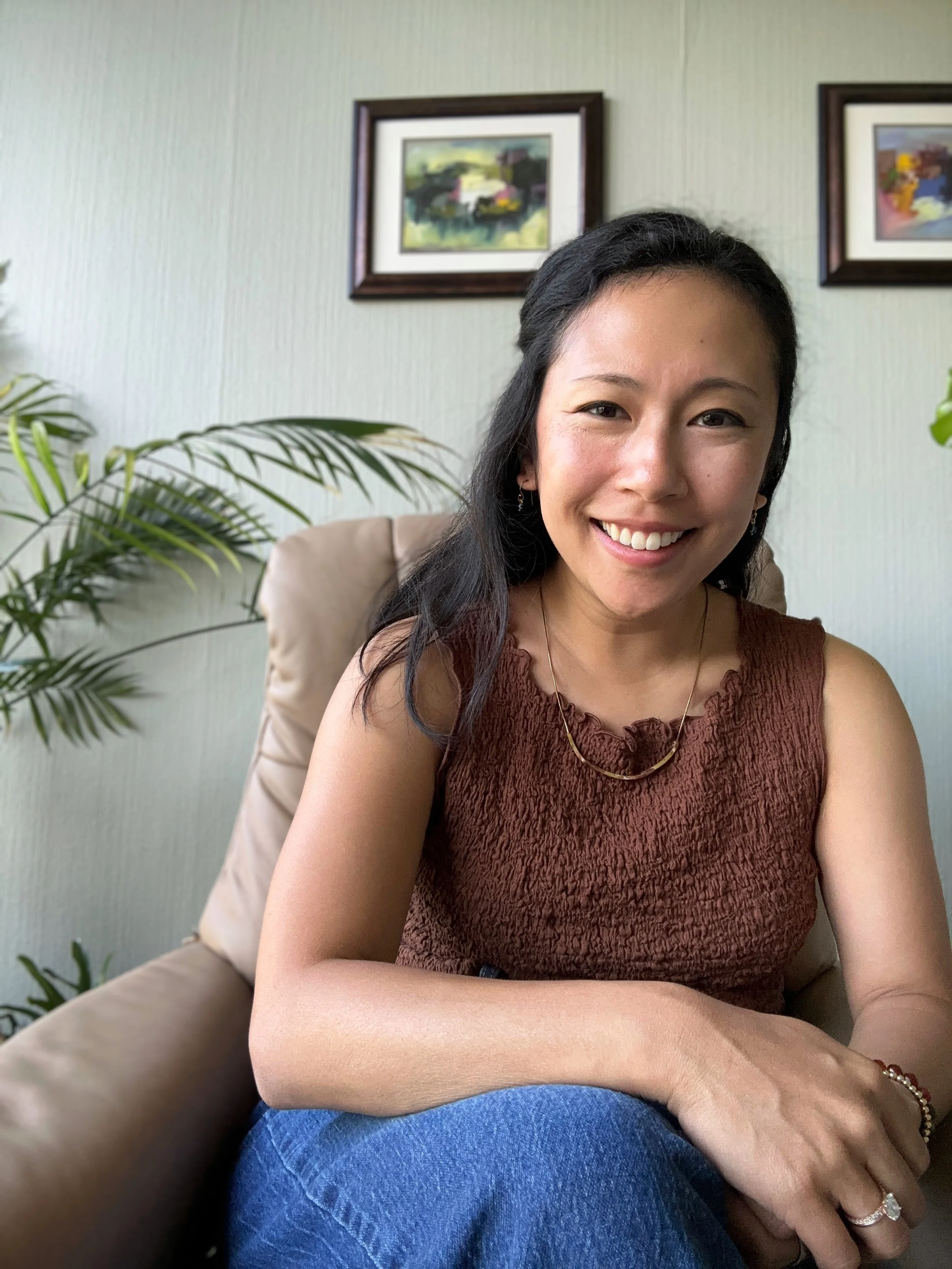 A woman smiling, sitting in a beige armchair with a wall decorated with framed paintings and a potted plant in the background.
