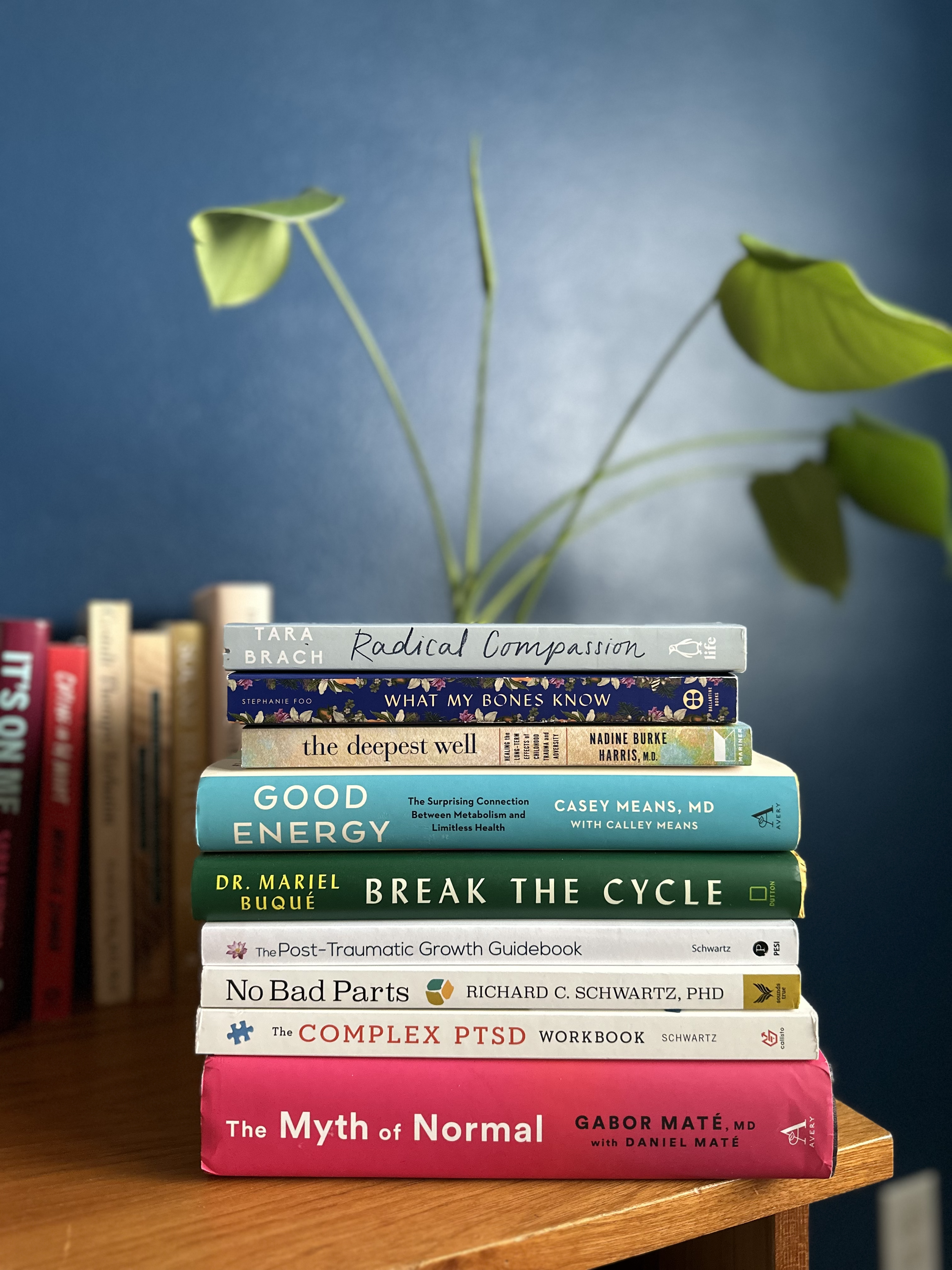 A stack of books on a wooden table with a blue wall background, including titles such as 'Radical Compassion', 'What My Bones Know', 'The Deepest Well', 'Good Energy', 'Break the Cycle', 'The Post-Traumatic Growth Guidebook', 'No Bad Parts', 'The Complex PTSD Workbook', and 'The Myth of Normal'.
