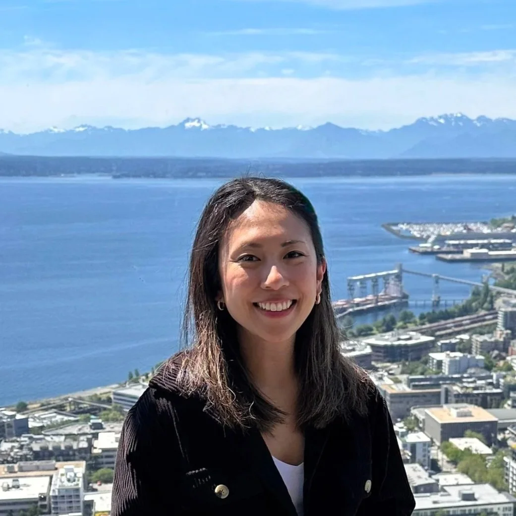 A woman smiling in front of a large body of water with mountains in the background and a cityscape below.